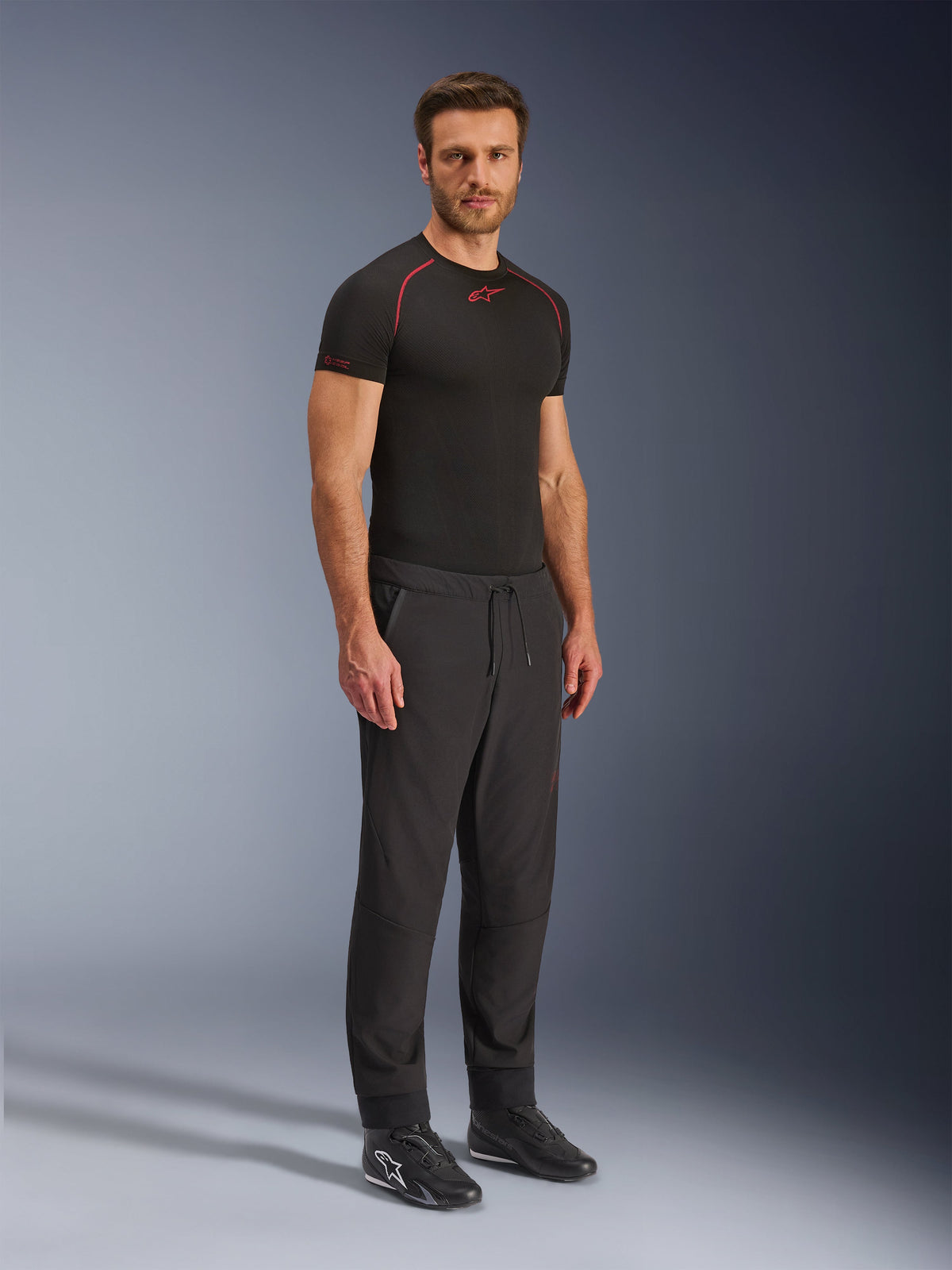 Alpinestars Aeroshell Airflow Pants, pantalon de moto noir, photo de studio en pied d'un mannequin portant un pantalon en softshell avec une taille à cordon de serrage et des chevilles élastiques, associé à un haut de sous-couche Alpinestars noir et des chaussures de moto noires.
