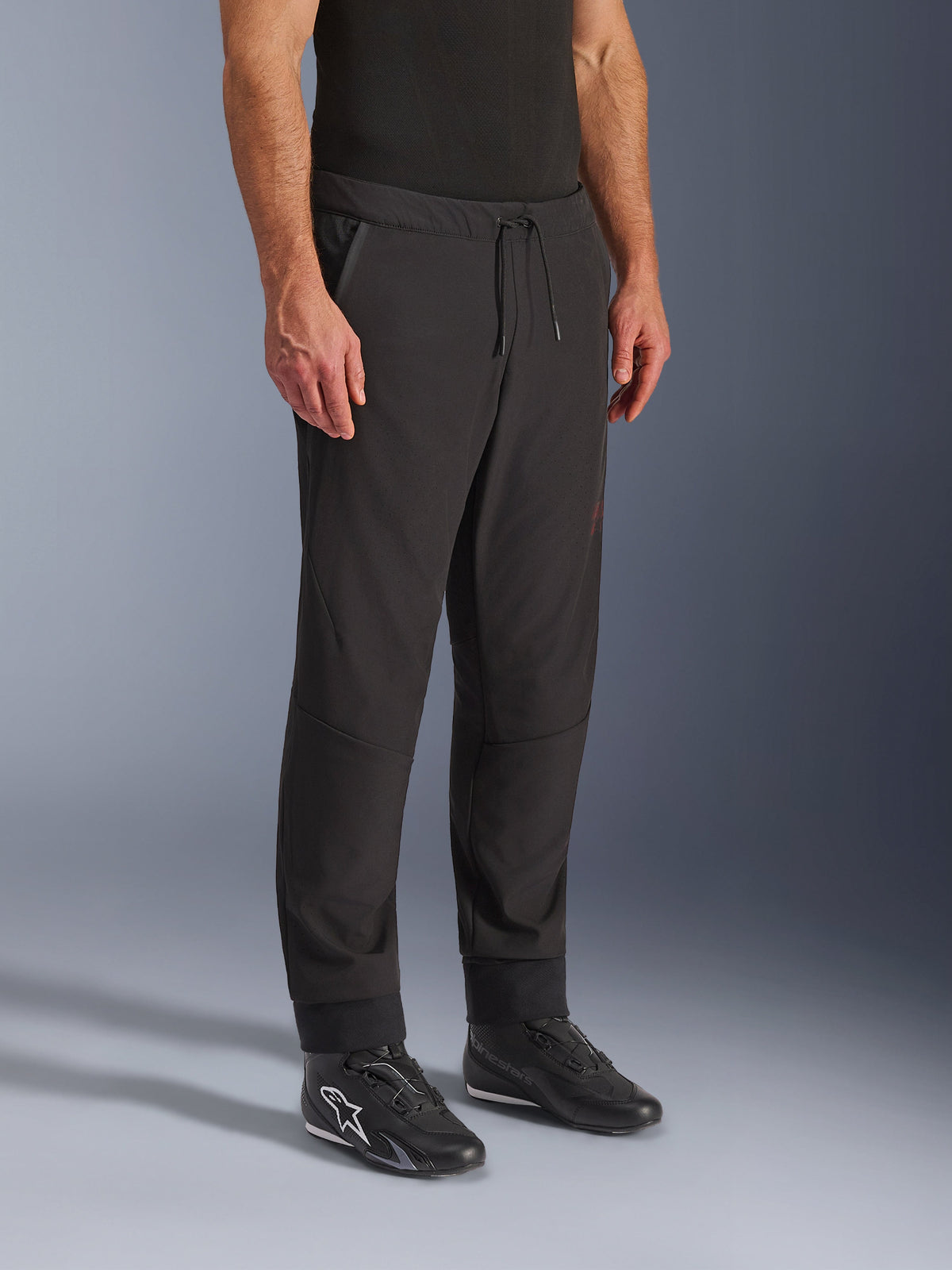 Pantalon Alpinestars Aeroshell Airflow, pantalon de moto noir, porté par un mannequin avec des chaussures de moto noires, doté d'une taille à cordon de serrage, de chevilles resserrées et d'un tissu softshell perforé au laser pour une ventilation maximale