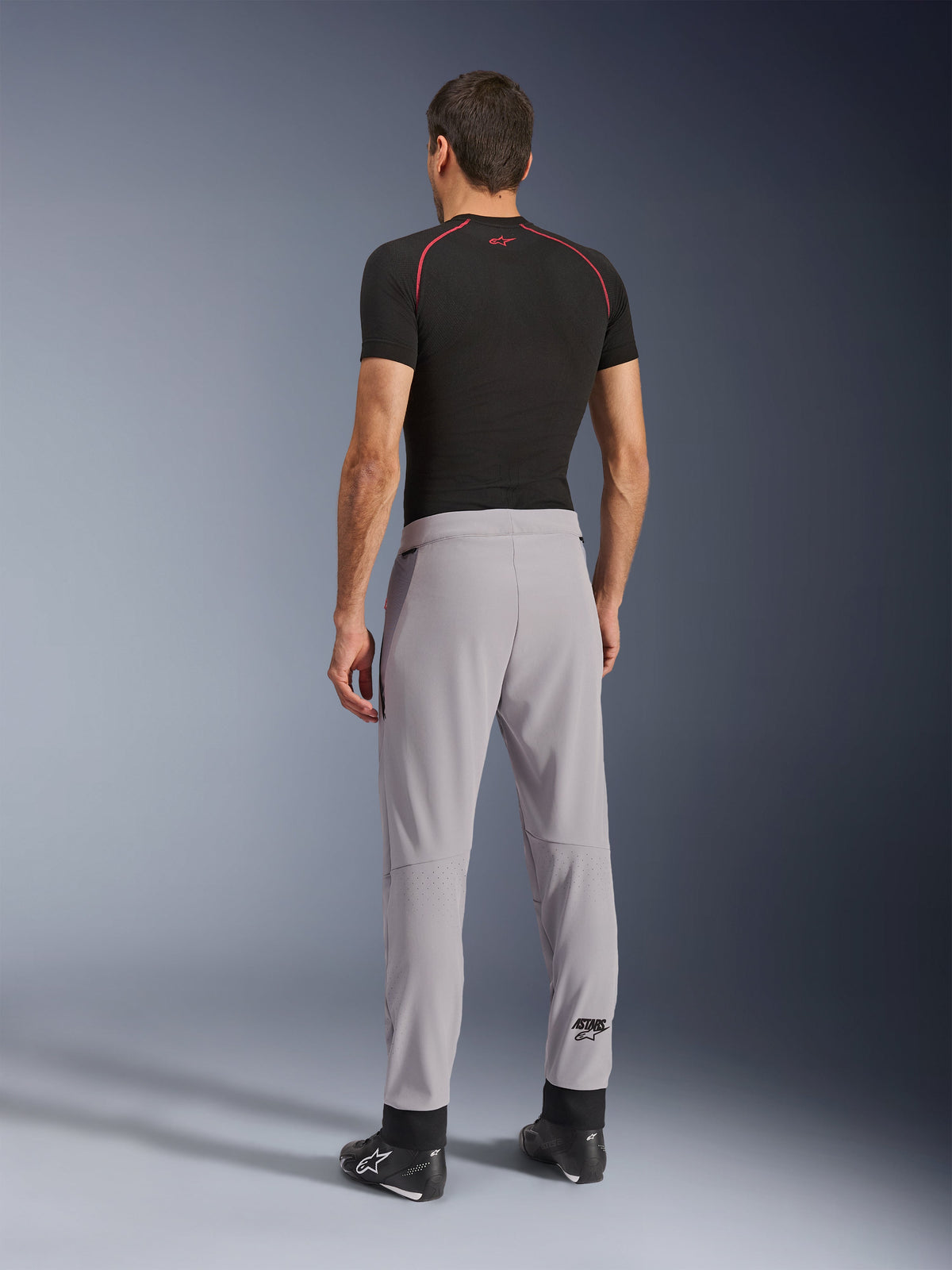 Rear view of a model wearing Alpinestars Aeroshell Airflow Pants in Mid Gray, paired with a black base layer shirt and black shoes, highlighting the laser-perforated zones on the calves and the athletic sport fit.