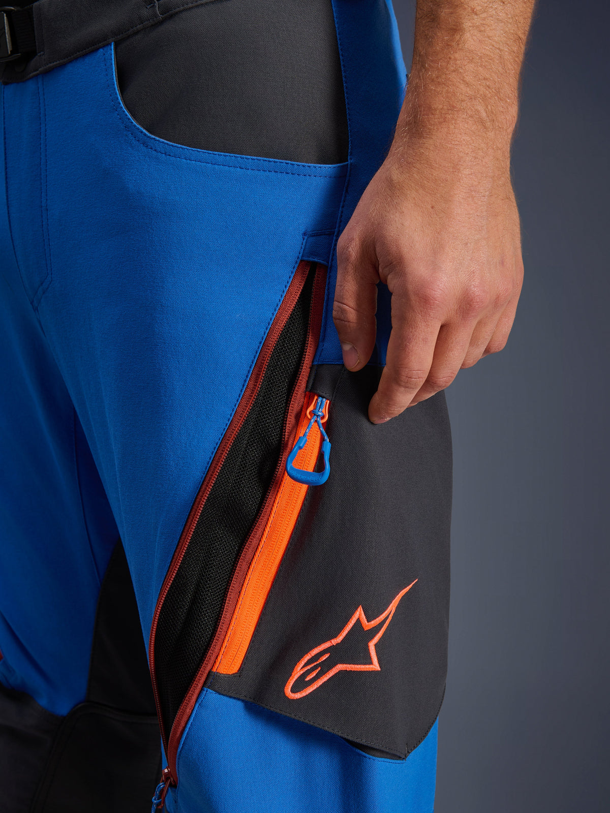 Alpinestars Maxdura Dual Pants detail, Royal Blue and Deep Gray adventure motorcycle pants, close-up of zippered thigh ventilation with orange accents and blue pull tab, orange Alpinestars logo on black panel, durable stretch polyamide construction