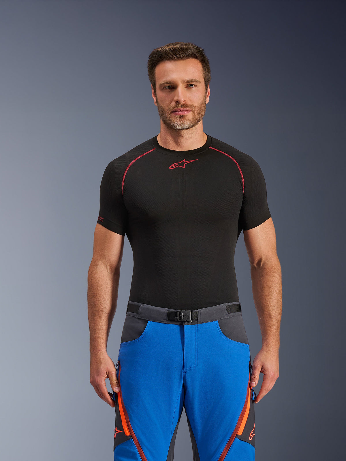 Alpinestars Maxdura Dual Pants, Royal Blue and Deep Gray, adventure motorcycle pants with orange accents, worn by model with a black compression shirt, featuring a magnetic buckle belt and zippered thigh vents, professional motorsport apparel