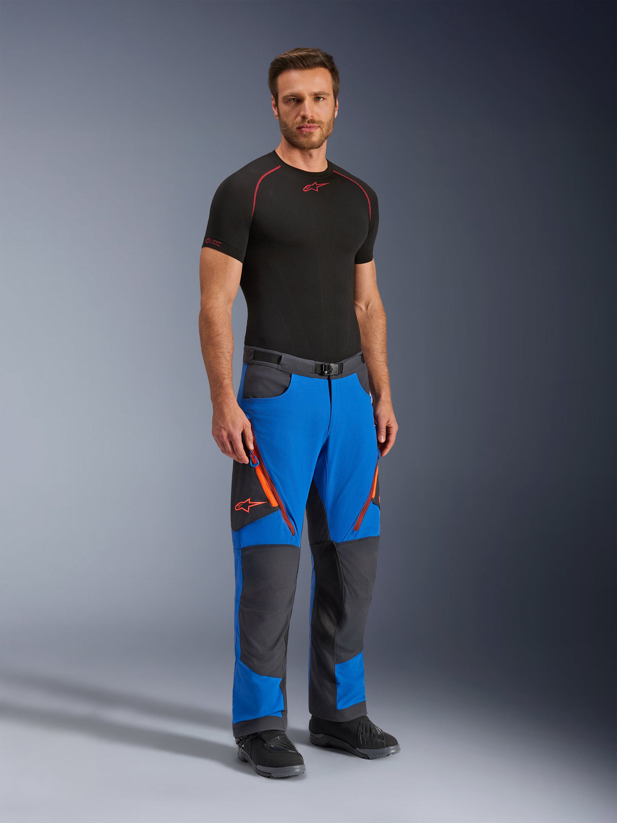 Alpinestars Maxdura Dual Pants, Adventure Motorcycle Pants, Royal Blue and Deep Gray, featuring orange zipper accents and logos, worn by a model with a black base layer and boots, technical design with reinforced panels for dual-sport riding