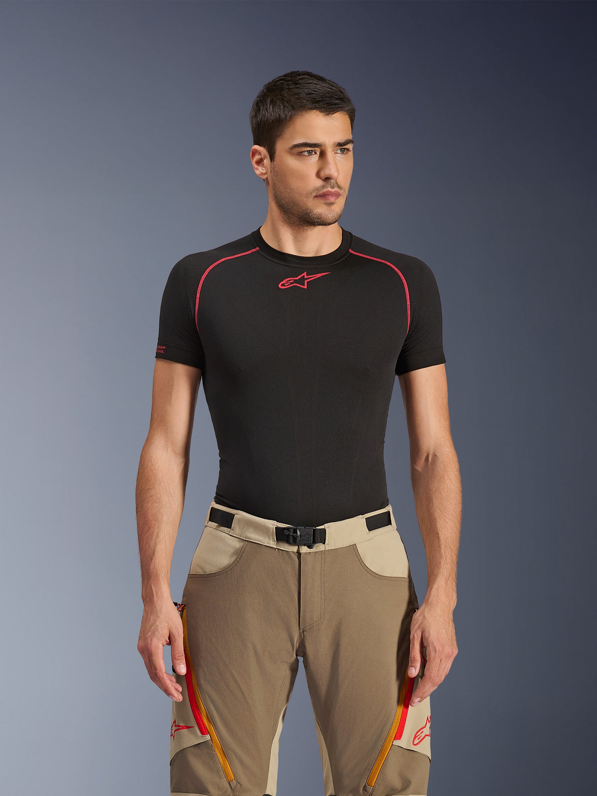 Alpinestars Maxdura Dual Pants in Walnut Aluminum, tan and beige adventure motorcycle pants worn by a model with a black base layer shirt, featuring orange zippered thigh vents, red branding, and a magnetic waist buckle, studio portrait.