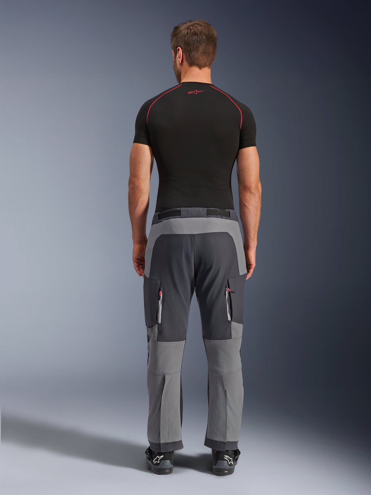 Pantalon Alpinestars Maxdura Dual, pantalon de moto d'aventure gris foncé et gris profond, vue de dos sur mannequin portant un t-shirt noir et des bottes, avec empiècements gris multi-tons et poches cargo zippées aux accents rouges.