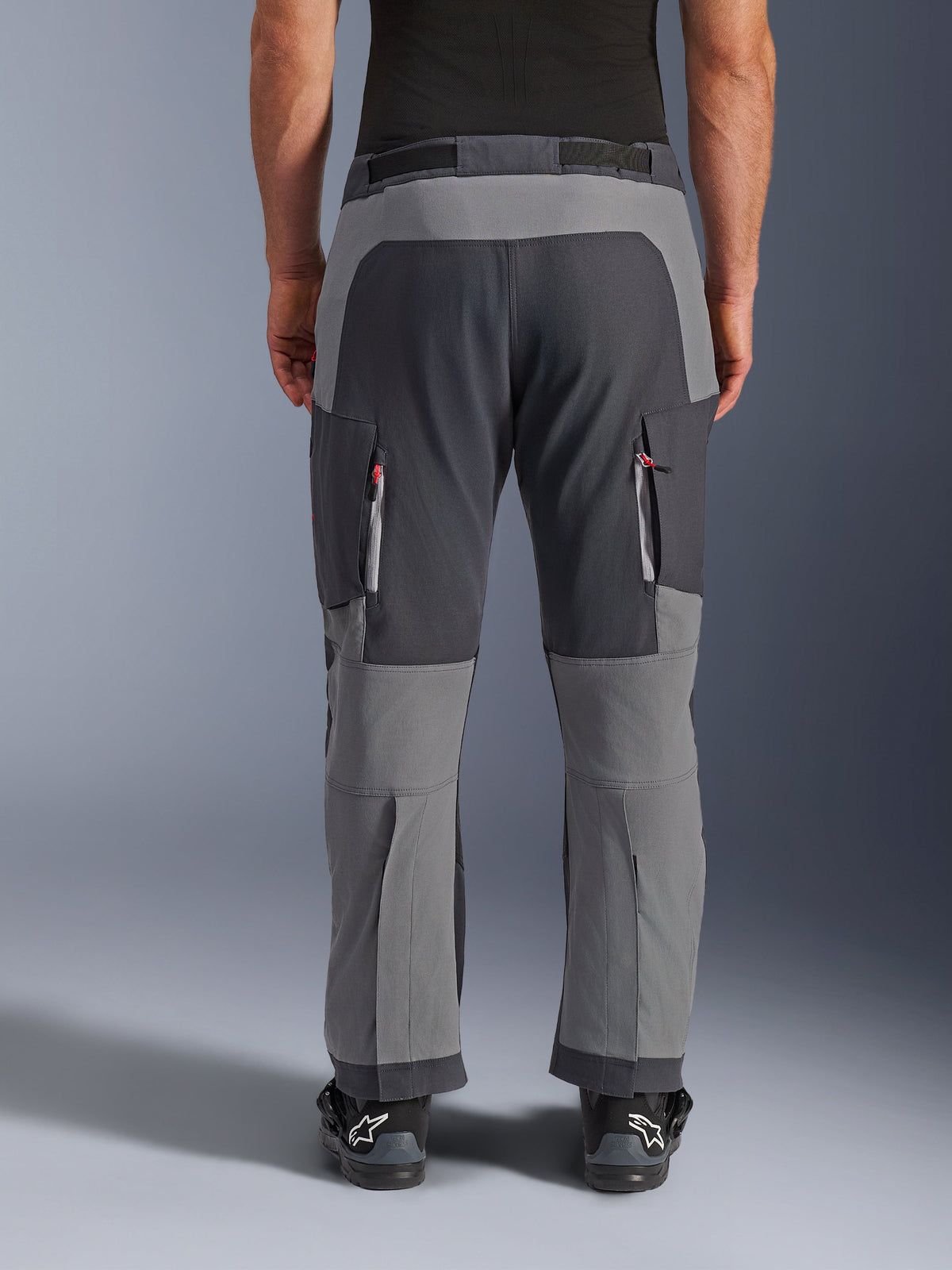 Alpinestars Maxdura Dual Pants, pantalon de moto aventure, gris foncé et gris profond, vue arrière soulignant les panneaux renforcés au niveau de l'assise et des genoux, ventilations zippées sur les cuisses, porté avec des bottes Alpinestars noires, construction robuste en polyamide extensible pour la pratique du dual-sport.