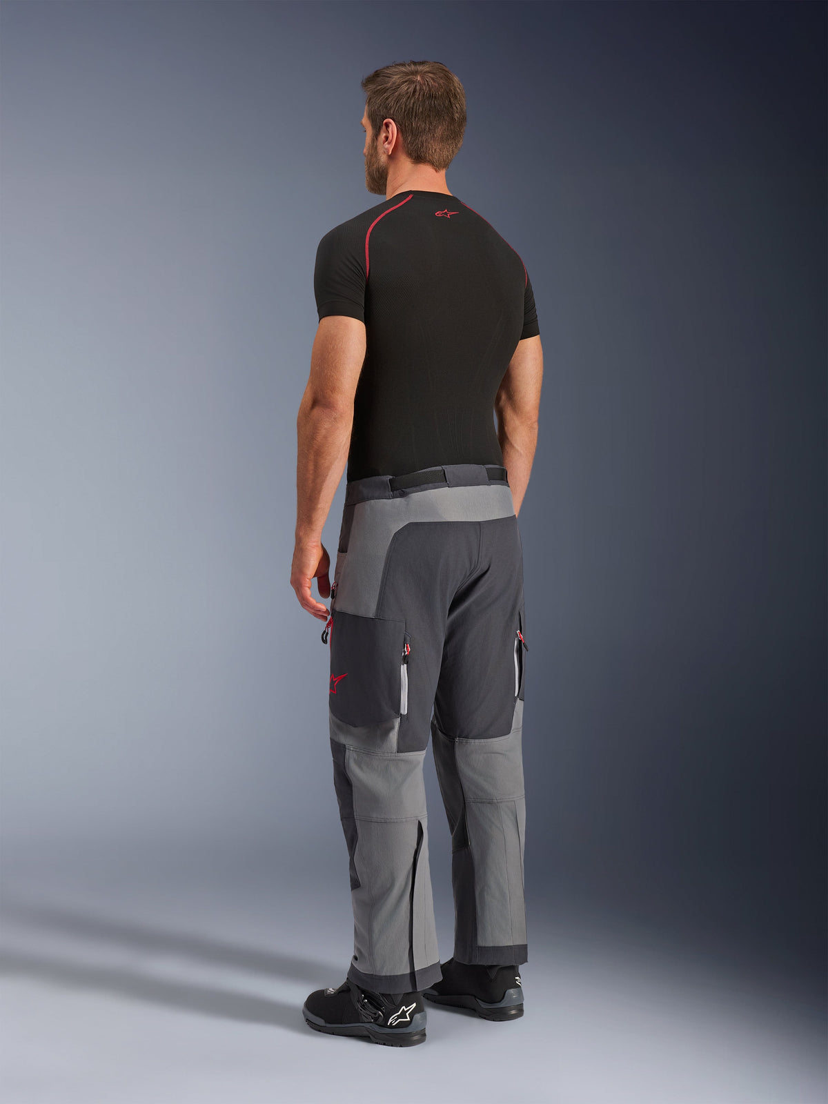 Alpinestars Maxdura Dual Pants, pantalon de moto d'aventure, gris foncé et gris anthracite, porté par un modèle de trois-quarts dos, avec des accents rouges sur les fermetures éclair et le logo, panneaux gris renforcés, poches cargo et coupe par-dessus les bottes