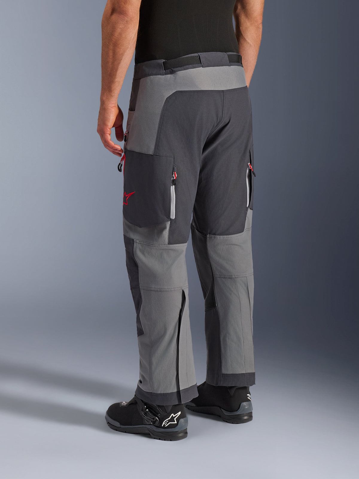 Alpinestars Maxdura Dual Pants, pantalon de moto d'aventure, gris foncé et gris profond, vue arrière présentant une construction multi-panneaux avec des touches de zips rouges et un logo étoile, associé à des bottes Alpinestars noires, conçu pour la performance dual-sport