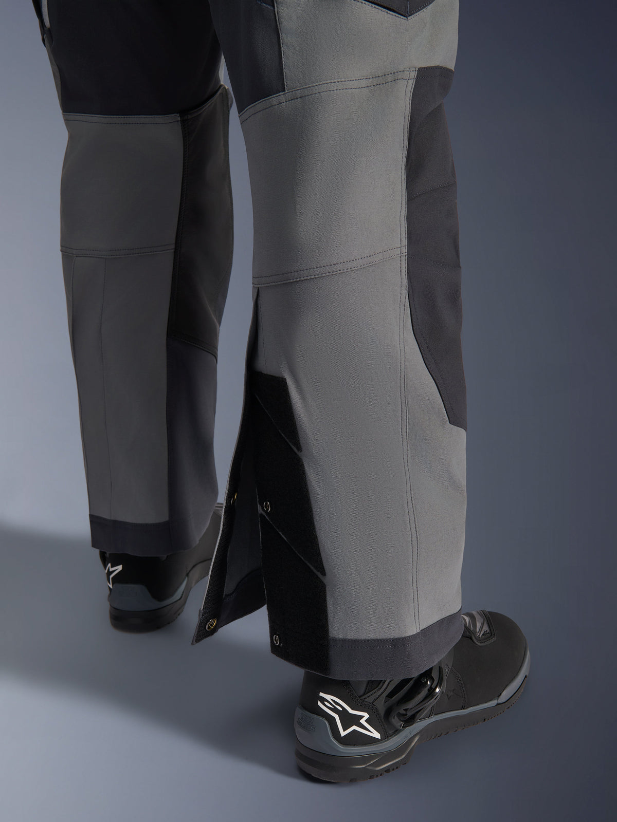 Alpinestars Maxdura Dual Pants, pantalon de moto aventure, gris foncé et gris profond, vue rapprochée du bas de jambe ajustable par-dessus la botte avec boutons-pression et panneaux renforcés, porté avec des bottes Alpinestars noires, design technique robuste pour le dual-sport