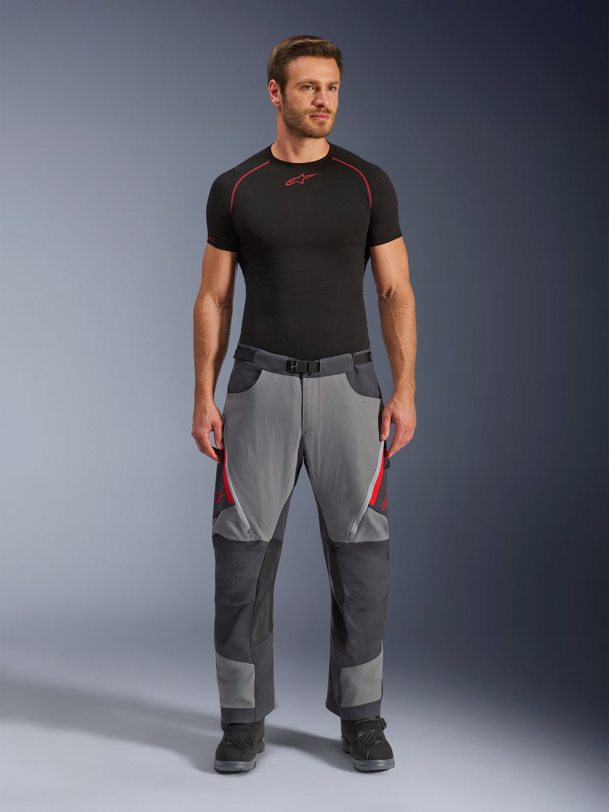 Alpinestars Maxdura Dual Pants, pantalon de moto, gris foncé et gris anthracite avec accents rouges, vue de face porté par un mannequin avec une couche de base Alpinestars noire et des bottes noires, doté de panneaux renforcés et d'aérations zippées sur les cuisses, équipement de pilotage de style aventure