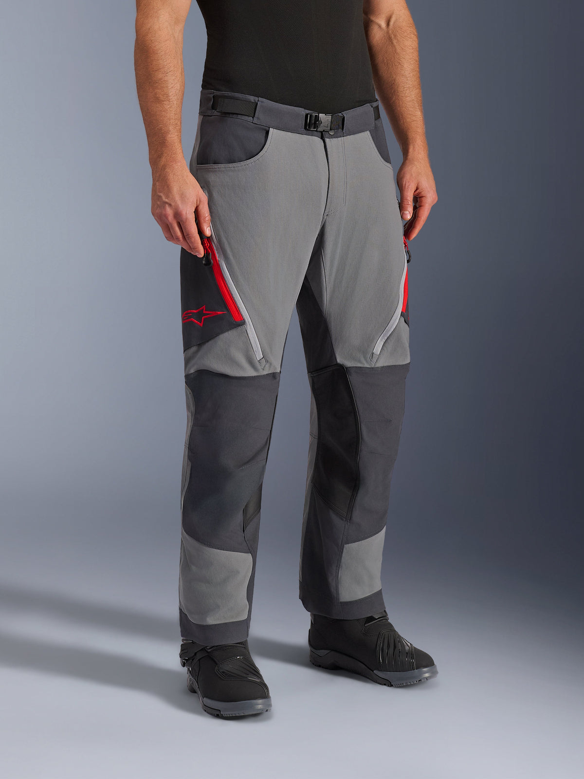 Pantalon Alpinestars Maxdura Dual en gris foncé et gris profond, pantalon de moto adventure-touring avec aérations zippées rouges sur les cuisses et logo étoile, empiècements sombres renforcés aux genoux, porté avec des bottes de moto noires
