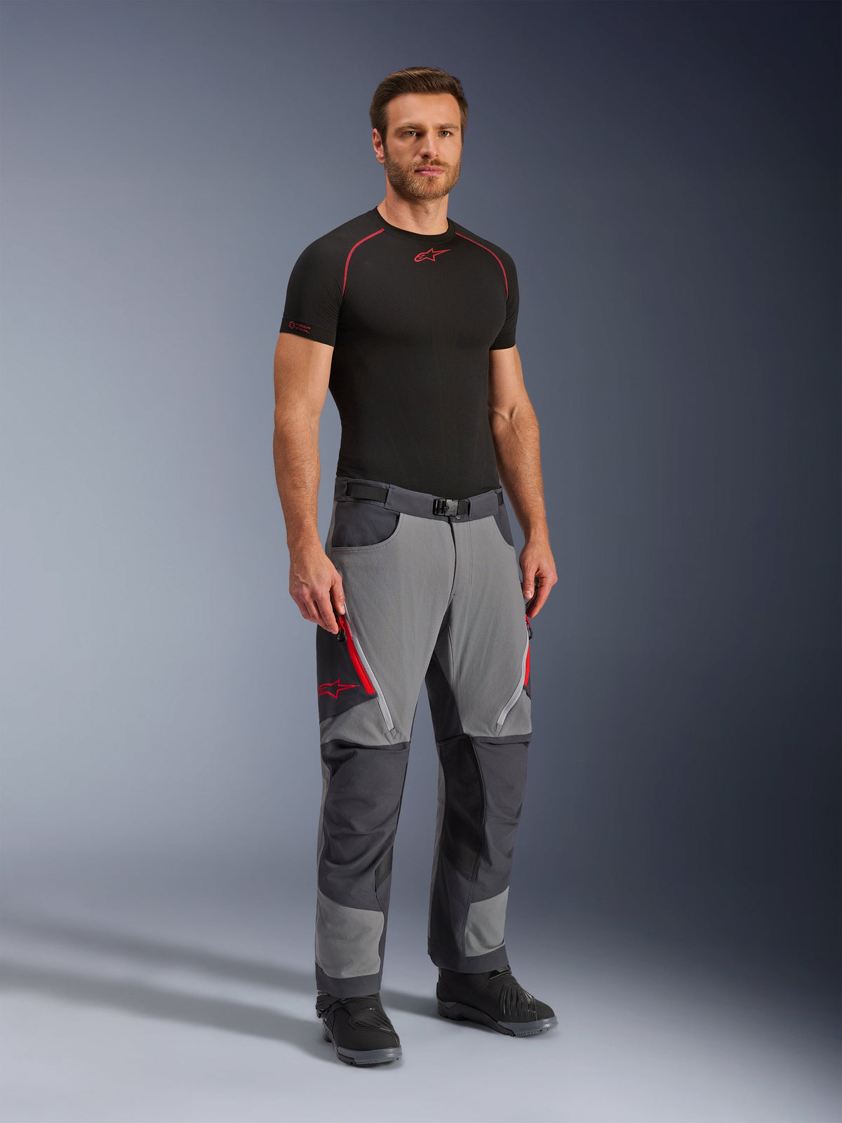 Pantalon Alpinestars Maxdura Dual, pantalon de moto aventure, gris foncé et gris anthracite, porté avec une couche de base noire et des bottes, doté d'aérations zippées rouges, de panneaux renforcés aux genoux et à l'assise, et d'une taille ceinturée, coupe athlétique pour la conduite dual-sport