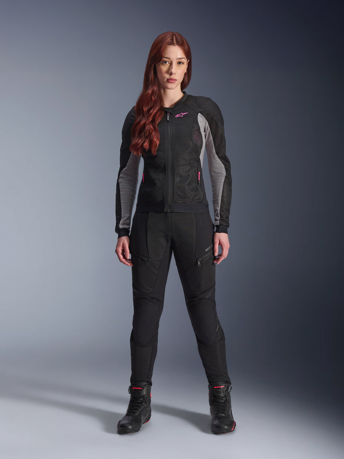 Veste et pantalon Alpinestars Stella Troop-Air, équipement de moto pour femme, noir avec accents gris cendre et rose fluo, porté par un mannequin aux cheveux roux, avec ventilation en mesh et design protecteur, pose debout en studio