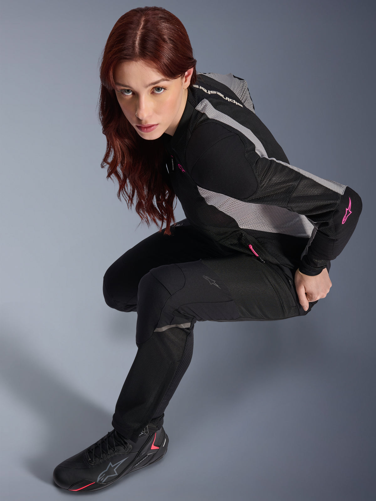 Femme portant le pantalon Alpinestars Stella Troop-Air, noir, pantalon technique léger en textile ventilé, associé à une veste en mesh noir et gris avec des touches de rose et des bottes de moto noires, portrait en studio, équipement de moto urbain