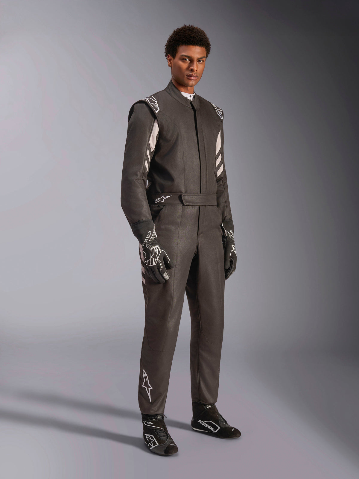 Pilote portant une combinaison Alpinestars GP Race V3 FIA, combinaison de course automobile noir et argent, avec gants et chaussures assortis, détails argentés sur les épaules et le bas des jambes, équipement de course automobile professionnel avec coupe ajustée