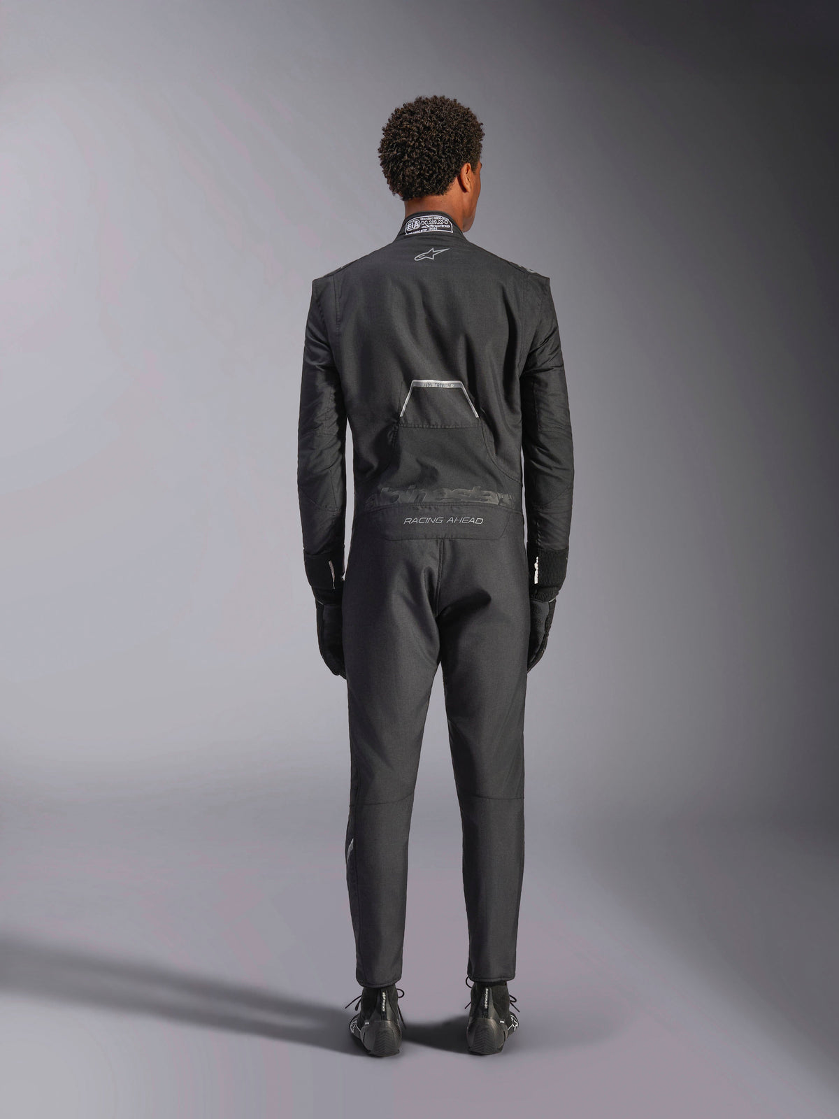 Combinaison Alpinestars Hypertech V3, Noir Black Dark Shadow, vue arrière d'une combinaison de course automobile professionnelle une pièce, arborant le label de certification FIA sur le col et les marquages 'Alpinestars' et 'RACING AHEAD' dans le bas du dos, portée avec gants et bottes de course noirs assortis.