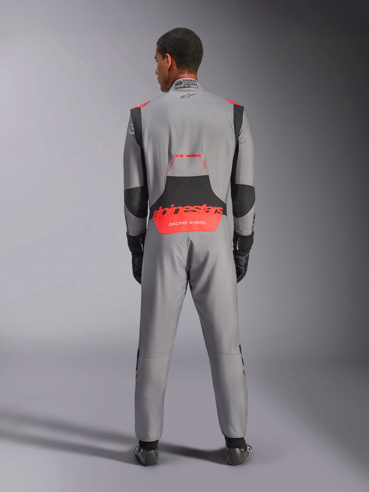 Combinaison Alpinestars Hypertech V3, combinaison de course automobile professionnelle, gris moyen avec accents noirs et rouge fluo, vue de dos sur un modèle avec logo Alpinestars rouge dans le bas du dos, construction légère en Nomex pour la performance sur piste