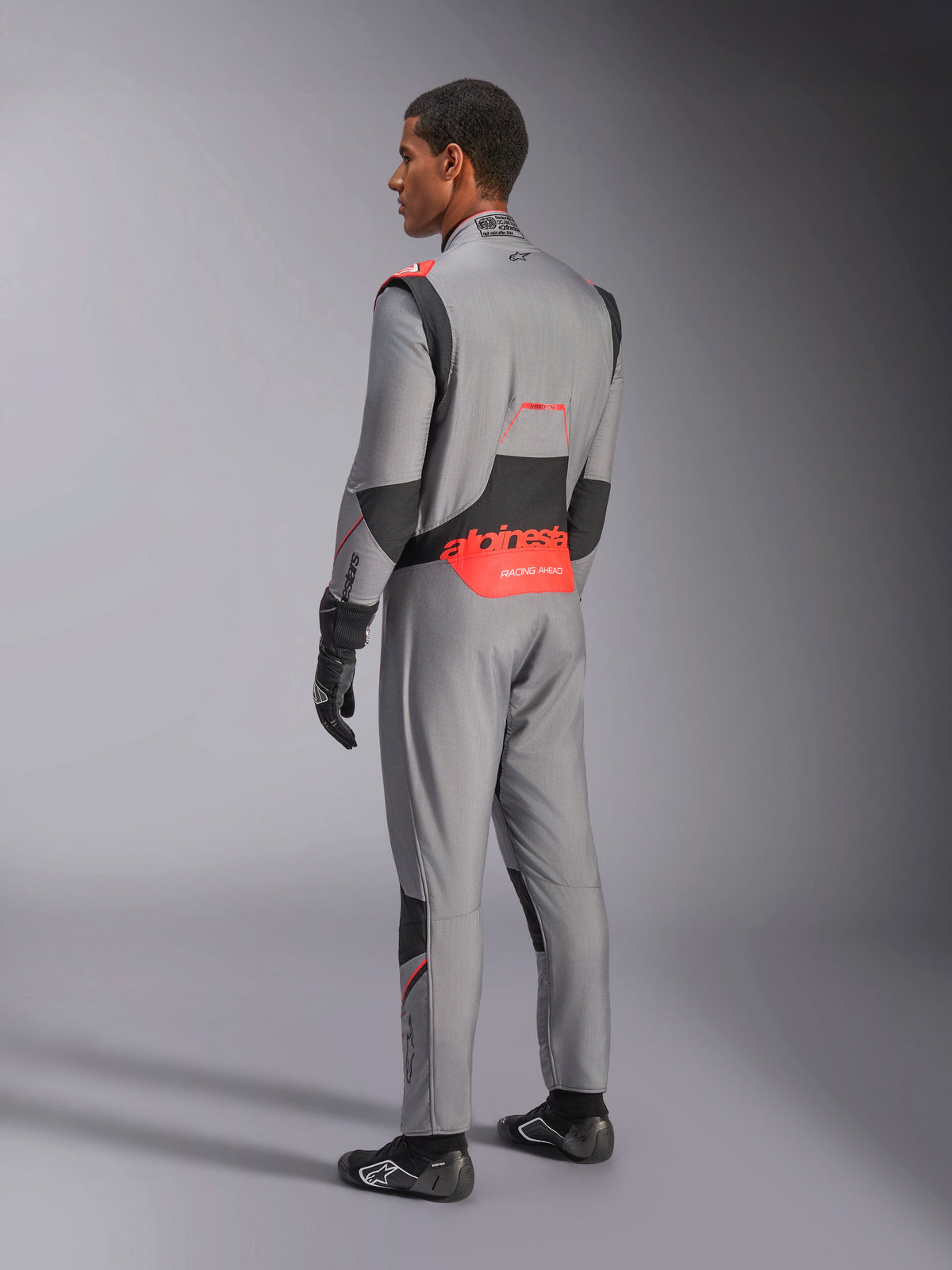 Combinaison Alpinestars Hypertech V3 en Gris Moyen Noir Rouge Fluo, vue arrière de trois-quarts, arborant le logo Alpinestars rouge sur le bas du dos, des panneaux extensibles noirs et une coupe athlétique pour la course automobile professionnelle.