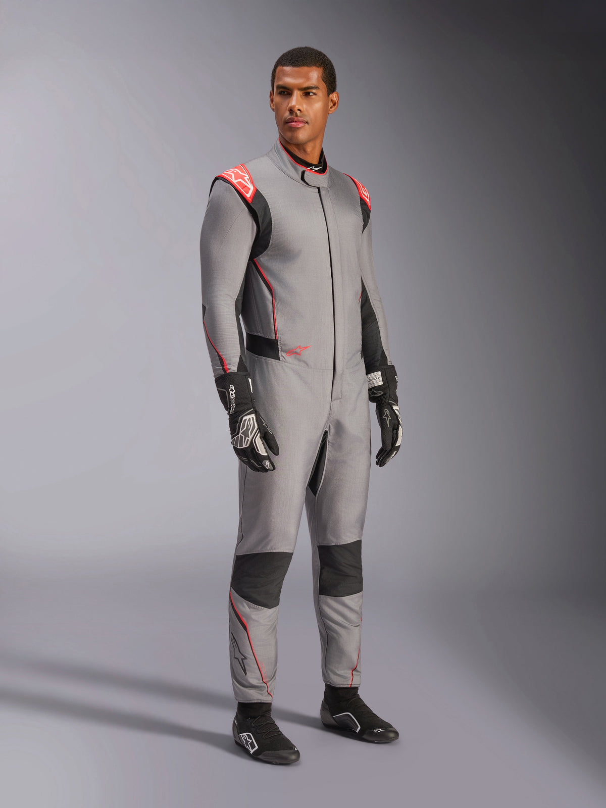 Modèle masculin portant la combinaison Alpinestars Hypertech V3 en gris moyen avec accents noir et rouge fluo, combinaison de course automobile professionnelle associée à des gants et des chaussures de course noirs, dotée d'une construction légère en Nomex® à deux couches avec des détails rouges aux épaules et une coupe athlétique