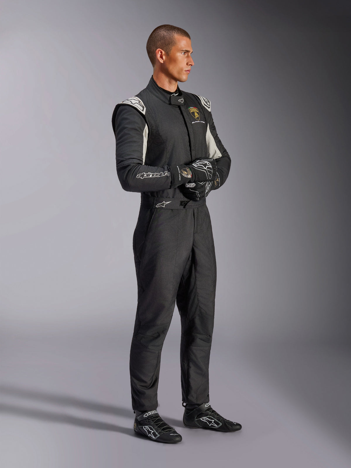 Male driver wearing the Alpinestars Limited Edition Lamborghini GP Tech V4 Suit, FIA-certified auto racing suit in Black, Silver, and Gold, paired with matching racing gloves and shoes, featuring Lamborghini Squadra Corse branding, professional motorsport racing gear