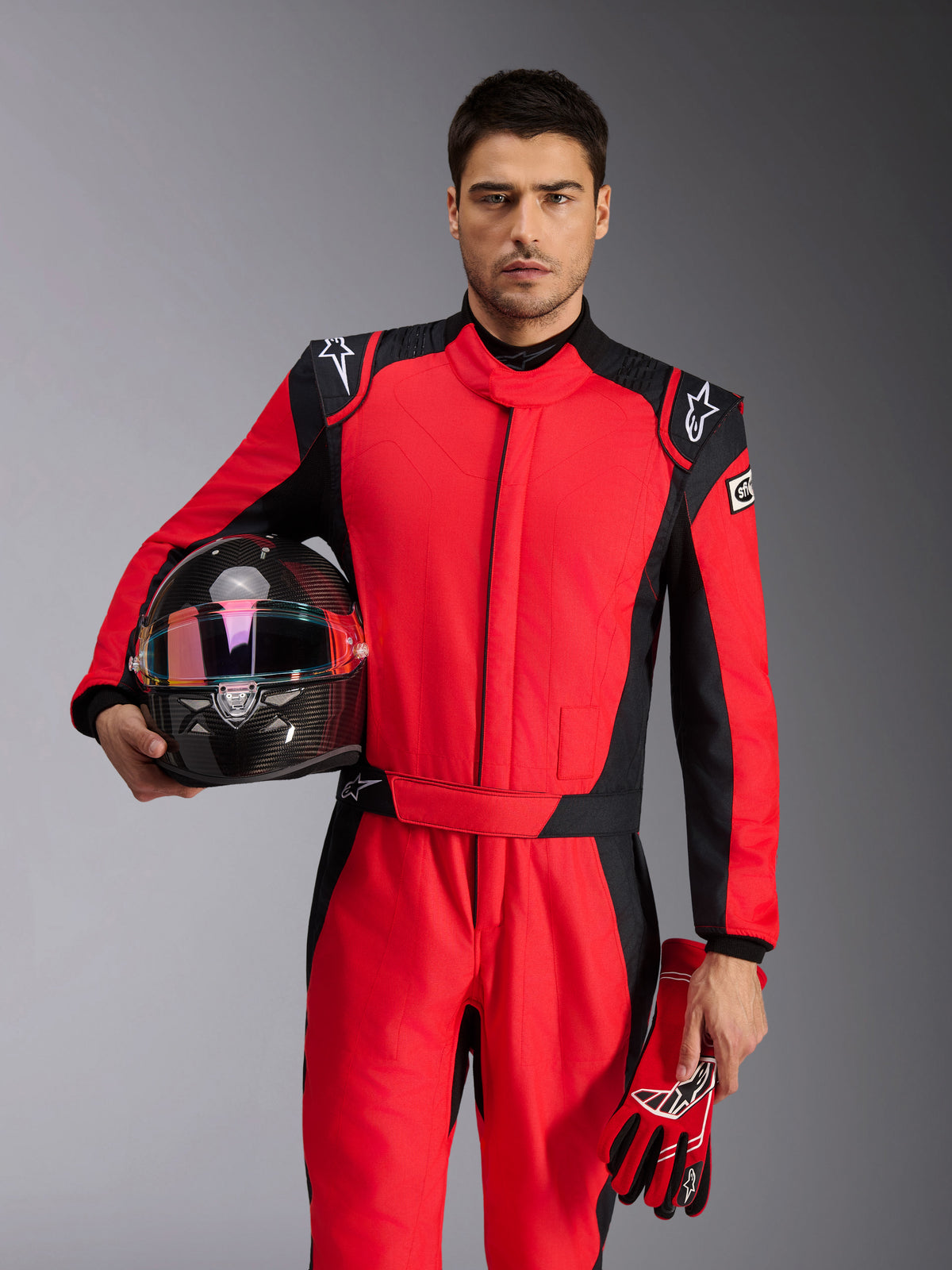 Pilote de course portant une combinaison Alpinestars GP Pro Comp V2 FIA rouge et noir, tenant un casque de course en fibre de carbone et des gants assortis, tenue de sport automobile professionnelle