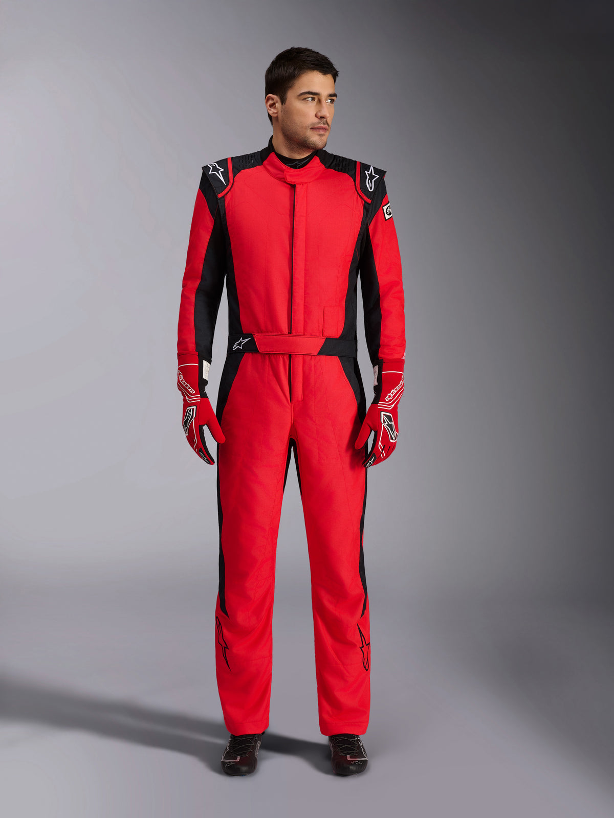 Modèle masculin portant la combinaison de course automobile Alpinestars GP Pro Comp V2 FIA, rouge et noir, associée à des gants de course rouges et des chaussures noires, combinaison de sport automobile professionnelle d'une seule pièce avec épaulettes et marquage Alpinestars, équipement de pilote haute performance pour la course automobile