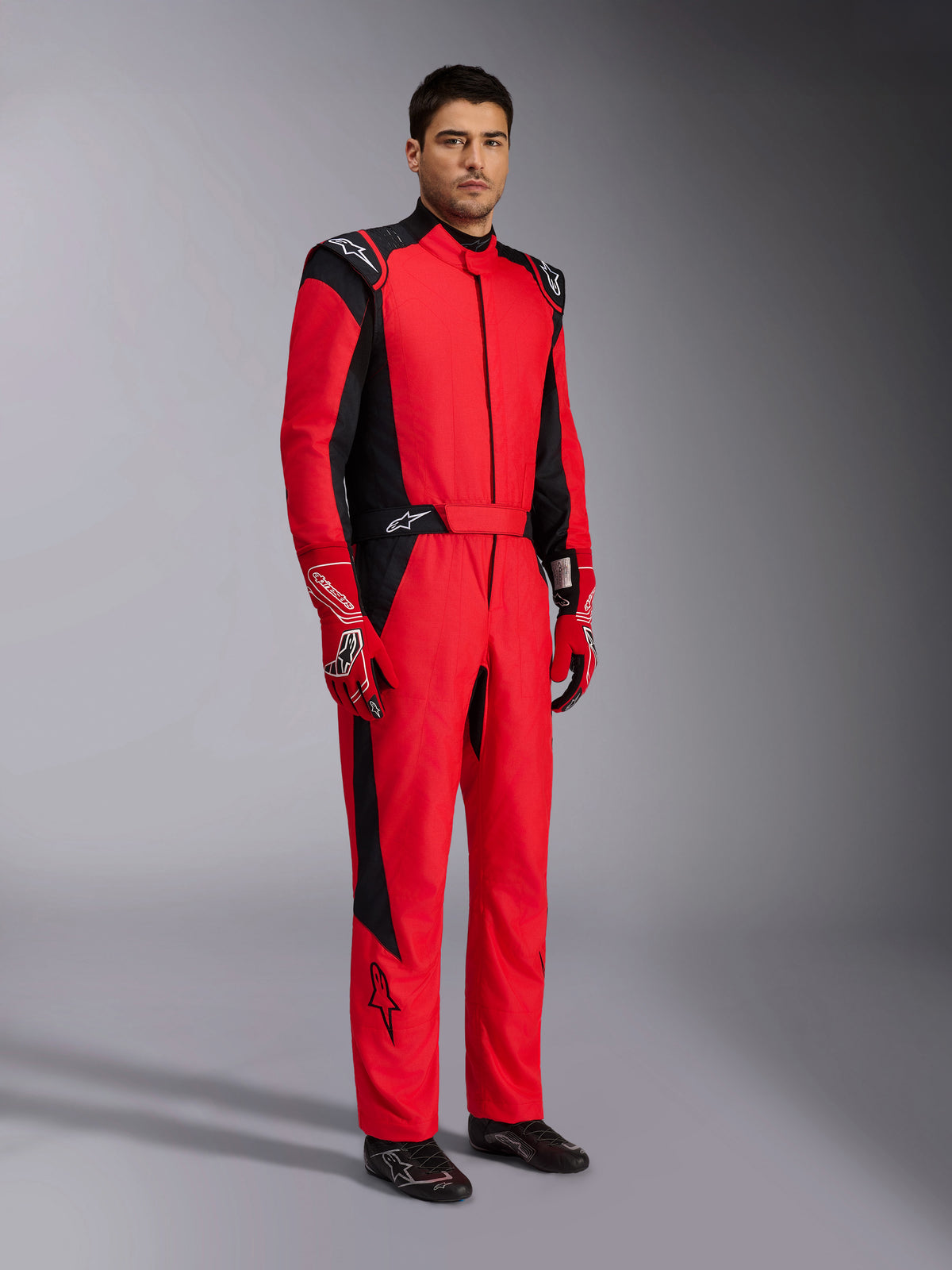 Combinaison de course Alpinestars GP Pro Comp V2 FIA, combinaison de course automobile professionnelle rouge et noire, portée avec des gants assortis rouges et blancs et des chaussures de course noires, portrait de studio en pied d'un équipement de protection pour sport automobile.