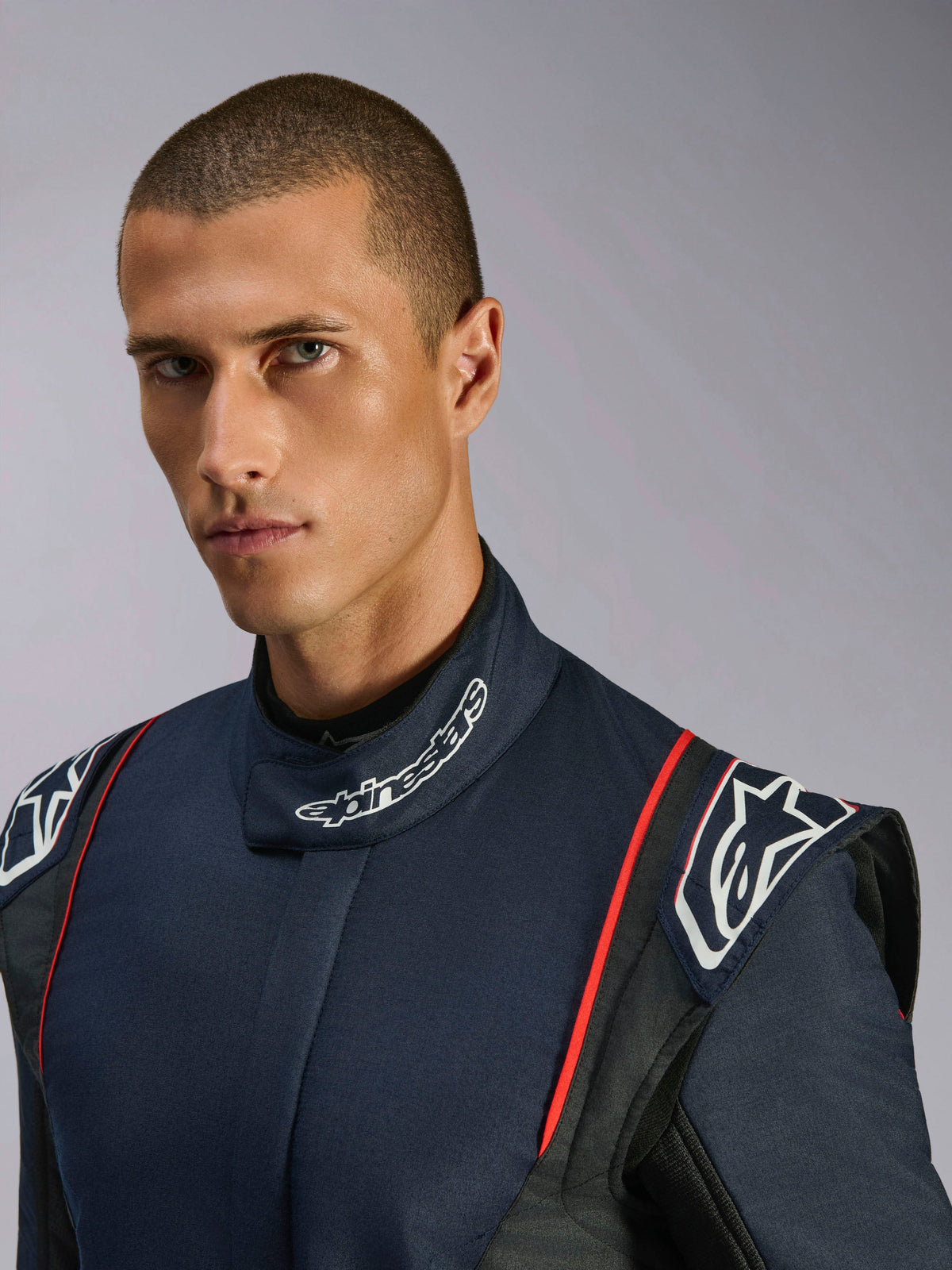 Close-up portrait of a model wearing the Alpinestars GP Tech V4 Suit, Auto Racing Suit in Blue Navy Black and Red, featuring Alpinestars branding on the high collar and structured shoulder epaulettes.