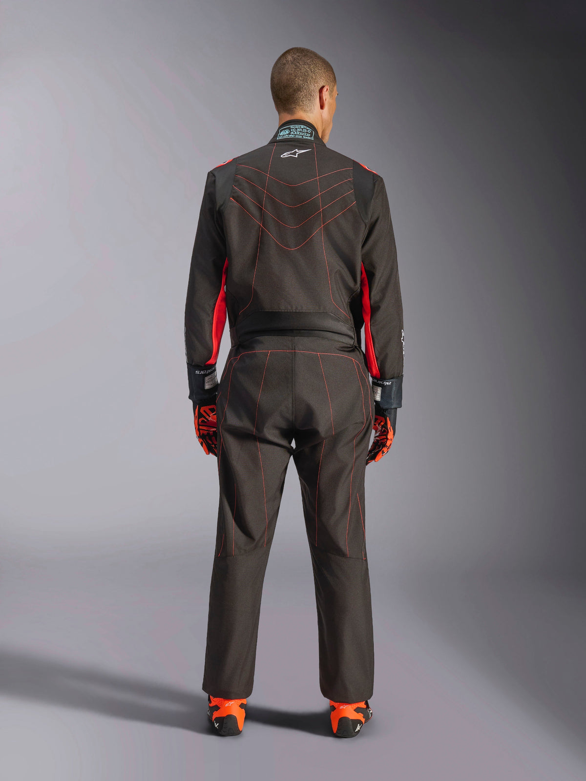 Vue arrière d'un pilote portant la combinaison Alpinestars KMX-9 V3, combinaison de karting, noire avec accents rouge fluo et surpiqûres contrastées rouges, équipement professionnel de sport automobile, portée avec des gants et des chaussures de course orange et noirs.