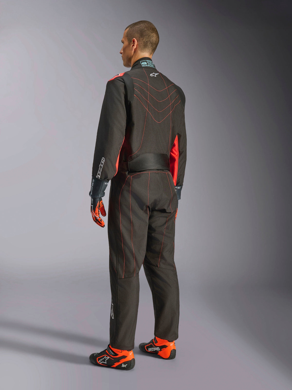 Combinaison Alpinestars KMX-9 V3, combinaison de karting, noir et rouge fluo, vue de dos, avec coutures contrastées rouges et panneaux latéraux fluorescents, portée avec des gants de course et des chaussures de karting, équipement de sport automobile professionnel