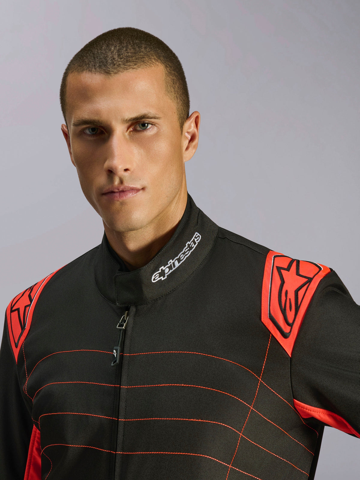 Gros plan d'un pilote portant la combinaison Alpinestars KMX-9 V3 en noir et rouge fluo, arborant un logo brodé sur le col et des épaulettes rouges avec le marquage Alpinestars, design d'équipement de karting technique