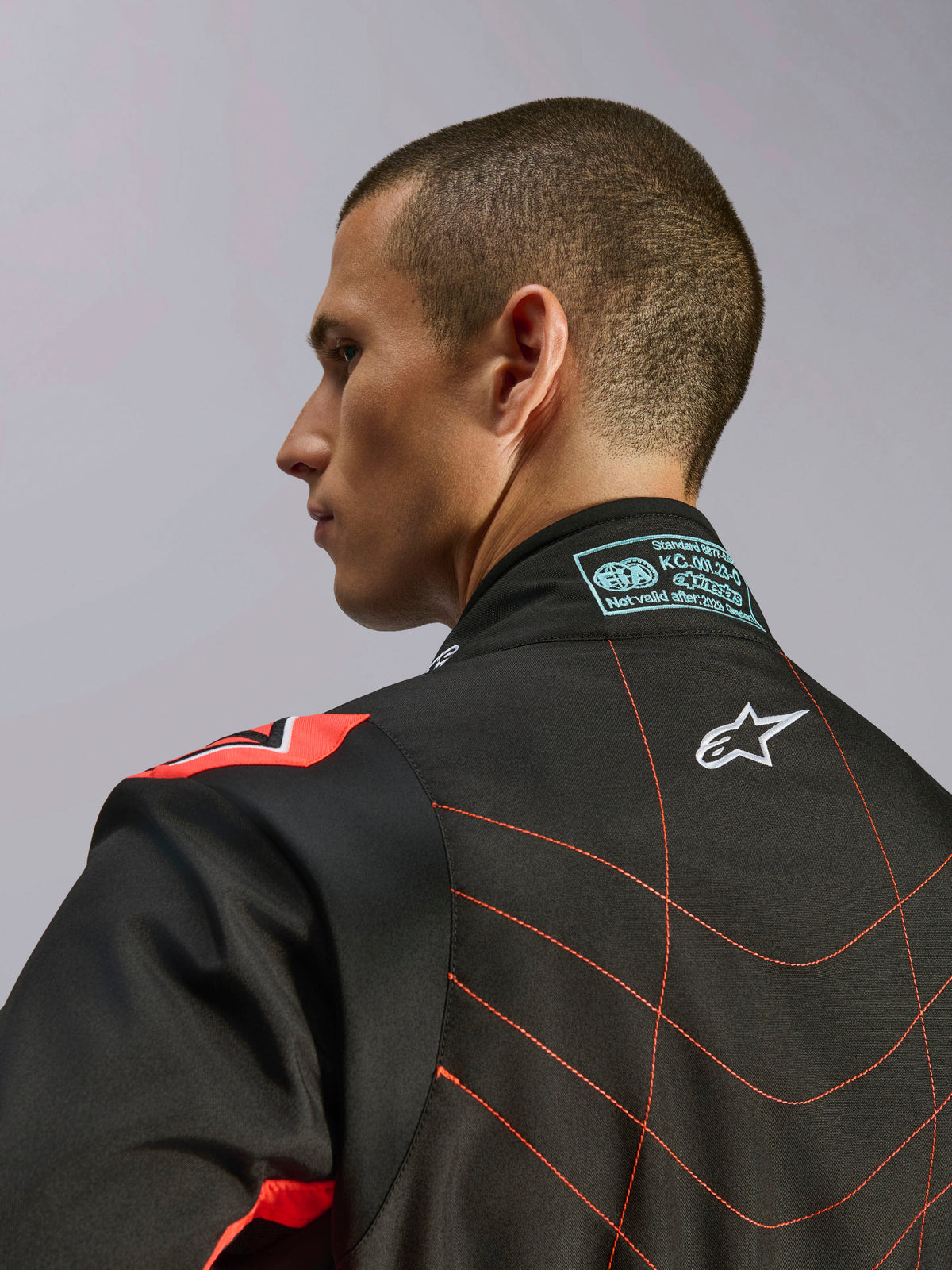 Combinaison Alpinestars KMX-9 V3, combinaison de karting noir et rouge fluo, vue rapprochée du haut du dos et du col sur un modèle, avec logo Alpinestars blanc, surpiqûres rouges détaillées et étiquette de certification FIA sur le col