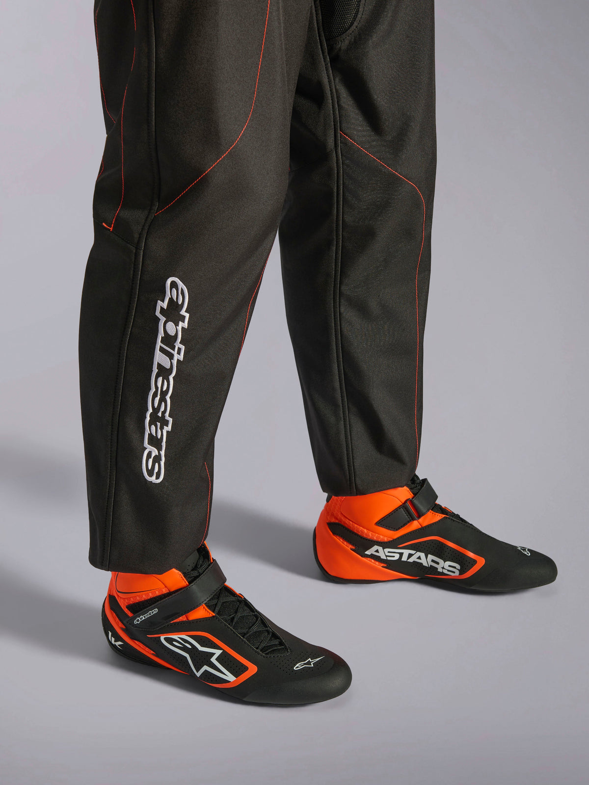 Combinaison Alpinestars KMX-9 V3, combinaison de karting, noir et rouge fluo, vue détaillée du bas des jambes et des chaussures de course, tissu noir avec surpiqûres rouge fluo, logo Alpinestars vertical sur le mollet, avec bottines de course noires et orange assorties, conçue pour le karting professionnel