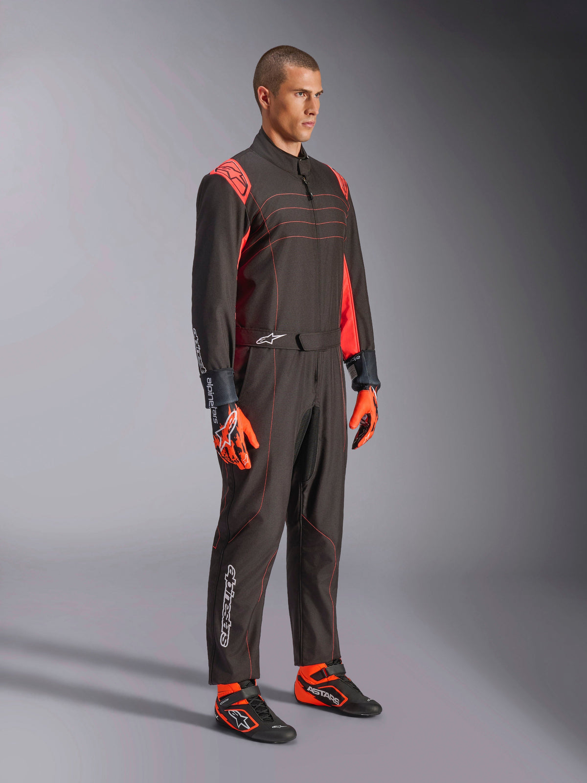 Combinaison Alpinestars KMX-9 V3, combinaison de karting, noir et rouge fluo, portée par un mannequin avec des gants orange et des chaussures de course noires, avec surpiqûres rouges, détails aux épaules et logos blancs, design athlétique pour la course professionnelle