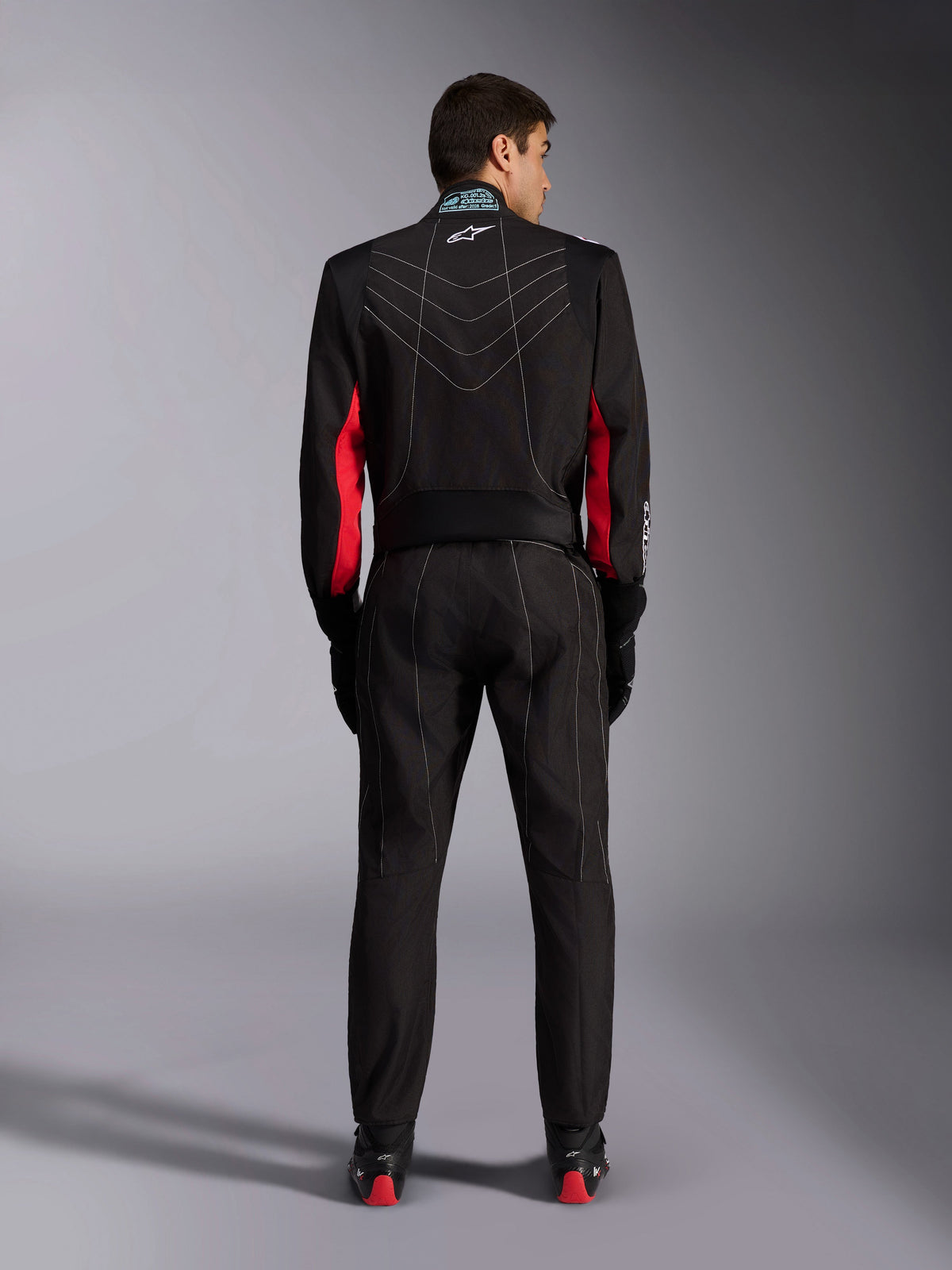 Combinaison Alpinestars KMX-9 V3, combinaison de karting, noire avec des accents rouges et blancs, vue de dos de la silhouette entière, présentant des surpiqûres contrastées blanches en forme de V, des empiècements rouges sur les manches et le logo Alpinestars sur le col, coordonnée avec des gants et des chaussures de course noirs