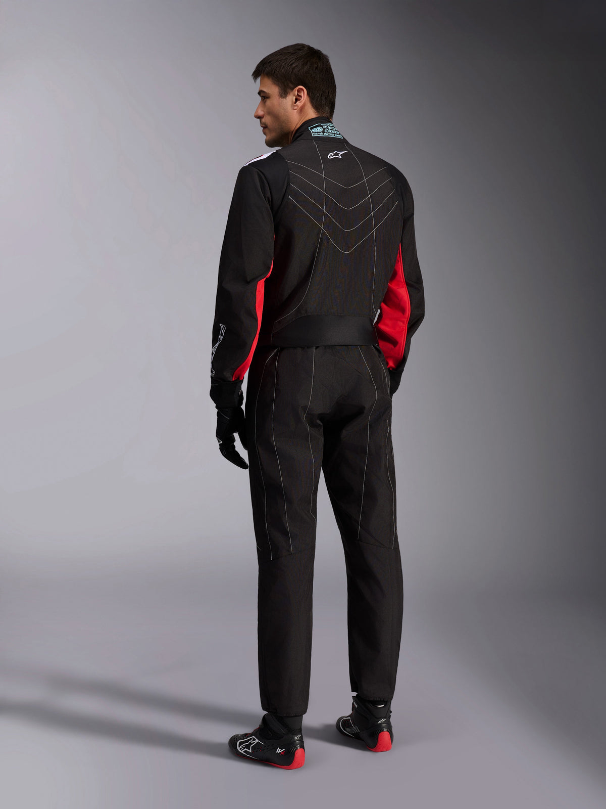 Combinaison Alpinestars KMX-9 V3, noir, rouge et blanc, combinaison de karting une pièce vue de dos, avec coutures géométriques blanches dans le dos et accents rouges sur les manches, portée avec des gants de course noirs et des chaussures de karting.