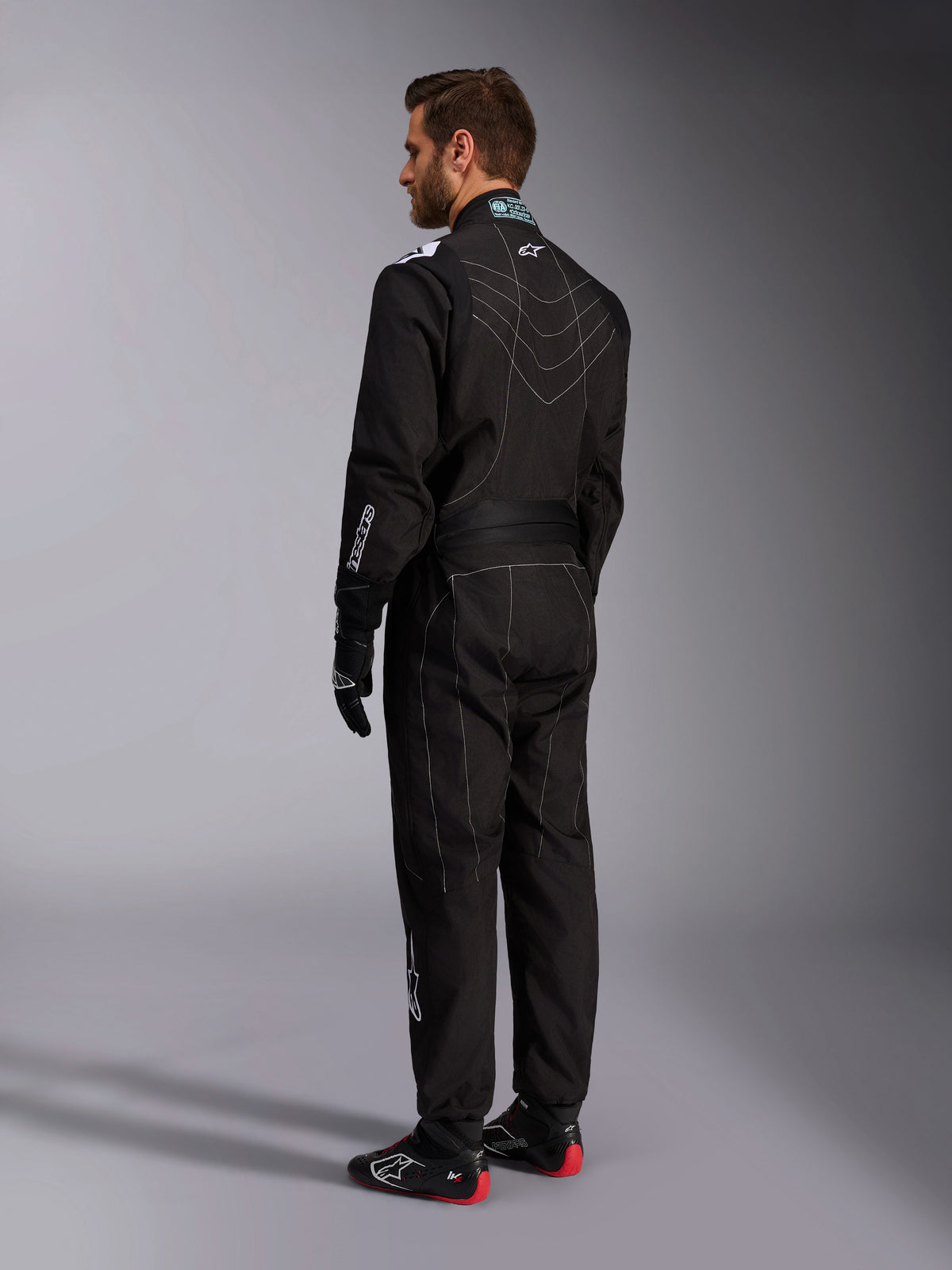 Combinaison Alpinestars KMX-9 V3, combinaison de karting noire et blanche, vue de trois-quarts arrière sur un mannequin homme, présentant des coutures contrastées blanches, des panneaux extensibles dans le dos et sur les épaules, des logos de marque blancs, portée avec des gants de course noirs et des chaussures noires à semelles rouges.