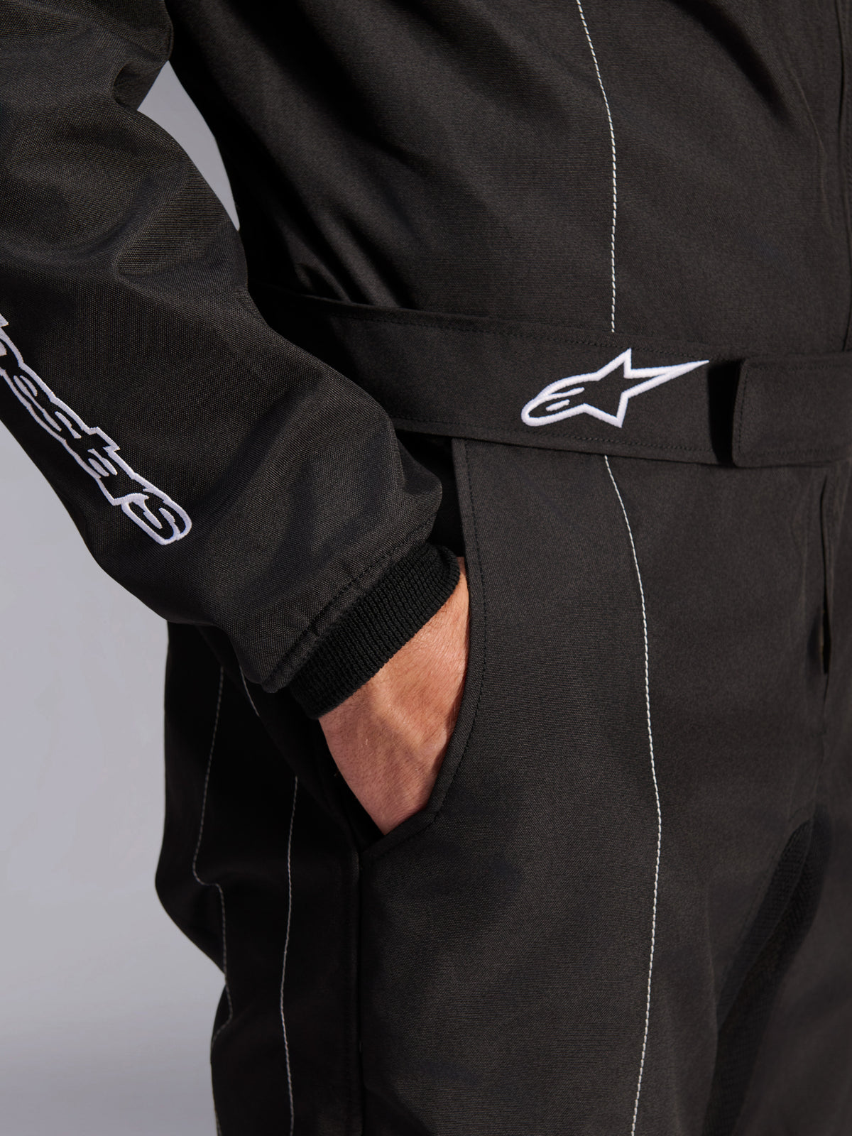 Combinaison Alpinestars KMX-9 V3, noir et blanc, vue rapprochée de la ceinture réglable avec logo brodé, poche latérale, surpiqûres blanches contrastées sur tissu noir, détail technique de combinaison de karting