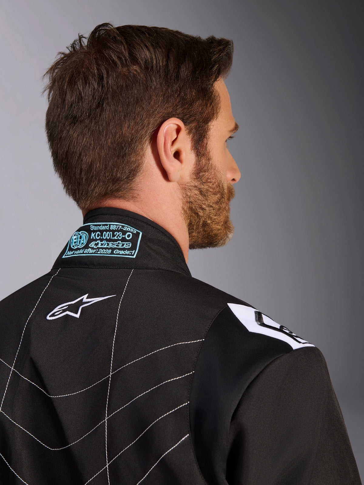 Combinaison Alpinestars KMX-9 V3, noir et blanc, gros plan sur le col arrière et la zone des épaules, présentant des détails d'homologation FIA brodés en bleu clair, un logo étoile blanc et des surpiqûres blanches contrastées.