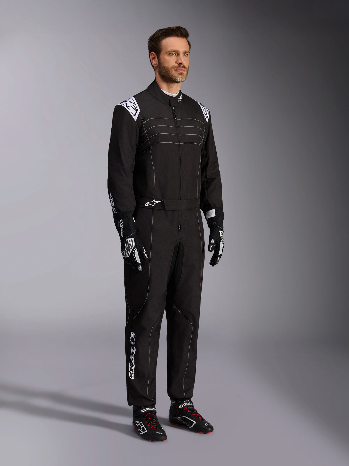Combinaison de karting Alpinestars KMX-9 V3, noir et blanc, mannequin homme portant une combinaison de course complète avec logo Alpinestars blanc sur les épaulettes, associée à des gants de course noirs et blancs et des chaussures de karting noires avec des lacets rouges, photo de studio montrant de face et de profil