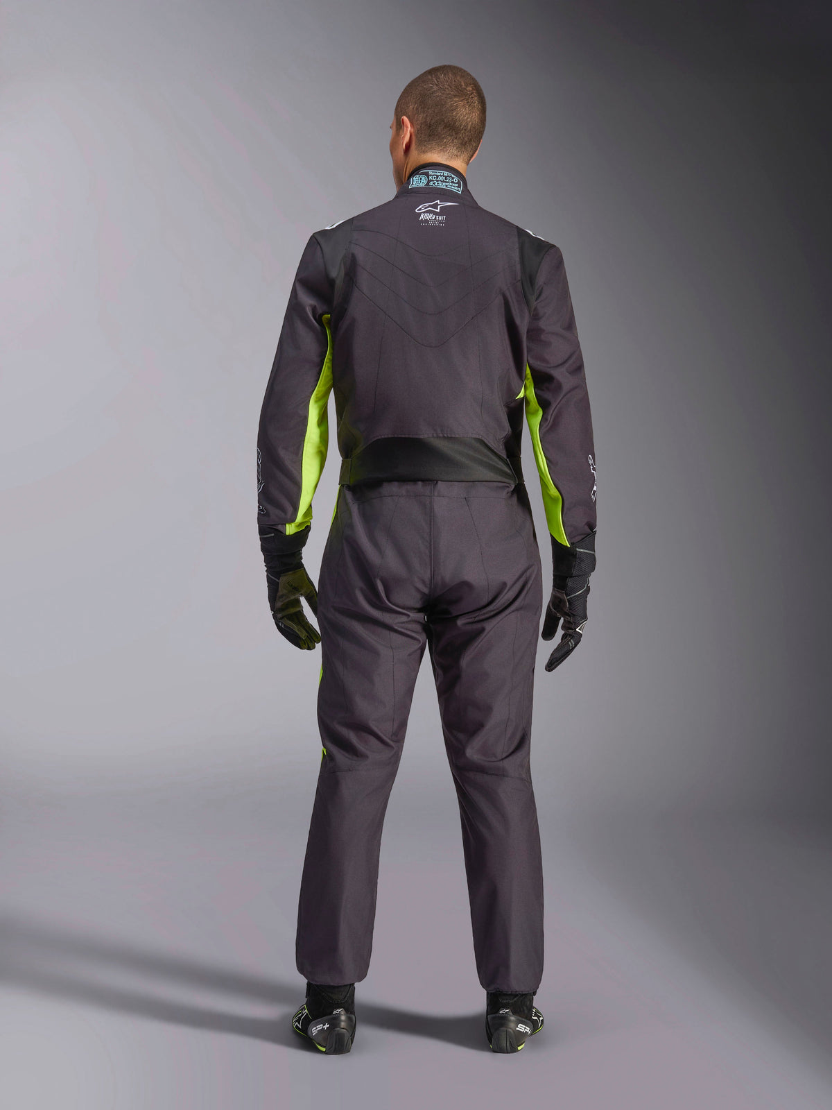 Vue arrière complète d'une personne portant la combinaison de karting Alpinestars KMX-9 V3 Graphic 3, coloris noir et jaune fluo, montrant les panneaux extensibles ergonomiques au dos et aux épaules, avec des gants de course et des chaussures noirs.