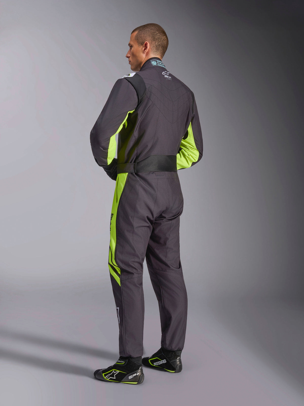 Combinaison Alpinestars KMX-9 V3 Graphic 3, combinaison de karting, noir et jaune fluo, vue arrière du modèle montrant la conception une pièce avec des accents jaune fluorescent sur les côtés et sous les bras, des panneaux extensibles ergonomiques sur la zone lombaire et une ceinture réglable à la taille, portée avec des chaussures de course assorties.