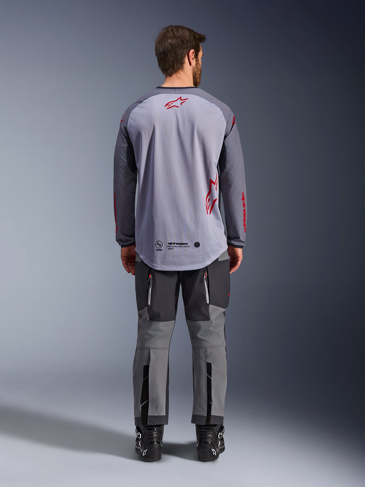 Male model from behind wearing the Alpinestars Maxdura Dual Jersey, Mid Gray and Dark Gray adventure jersey with red logo accents, mesh back construction, worn with matching gray off-road pants and black boots
