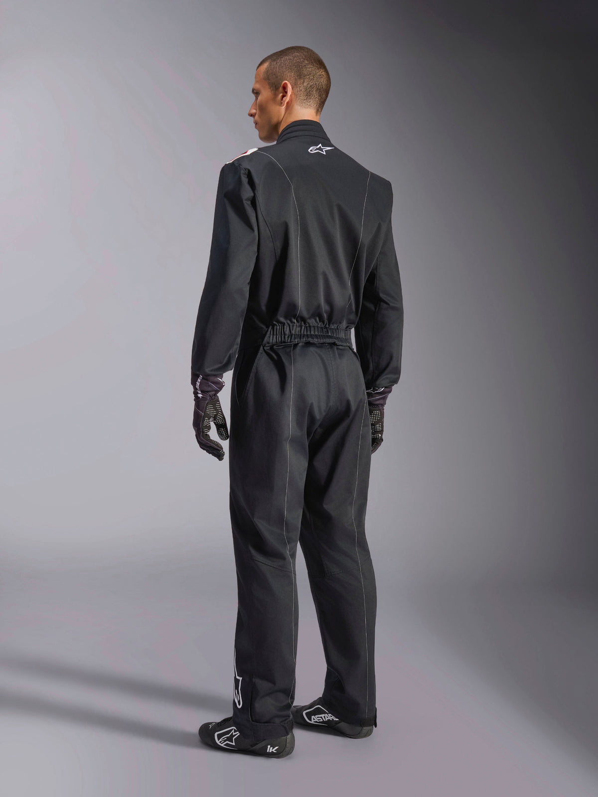 Alpinestars Kart Indoor V2 Suit, combinaison de karting une pièce noire présentée sous un angle de trois-quarts de dos, avec une taille élastique et un logo blanc sur le col, portée avec des gants et des chaussures de course noirs, conception durable pour le karting de loisir
