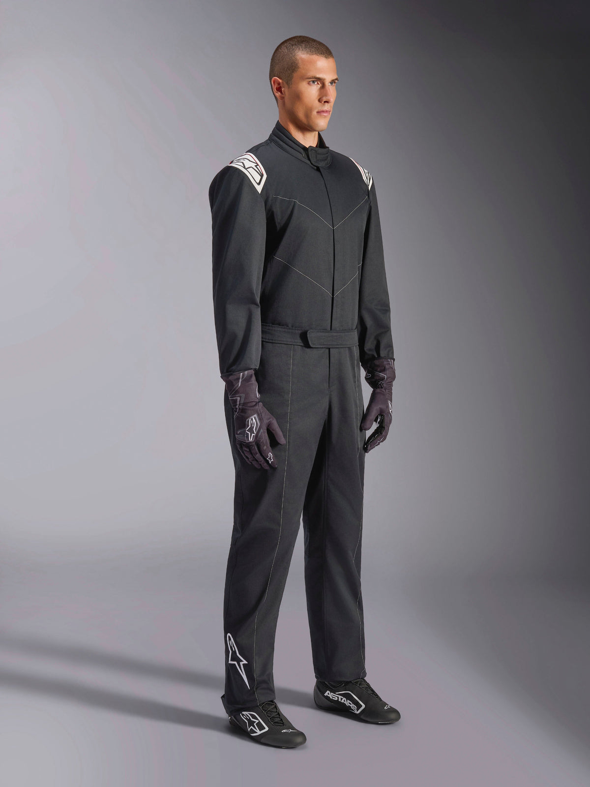 Combinaison Alpinestars Kart Indoor V2, combinaison de karting une pièce noire, portée par un mannequin avec des gants et des chaussures de course, dotée d'accents blancs aux épaules et d'un logo Astars blanc sur le bas de la jambe, vêtement de sport automobile professionnel pour le karting en intérieur.