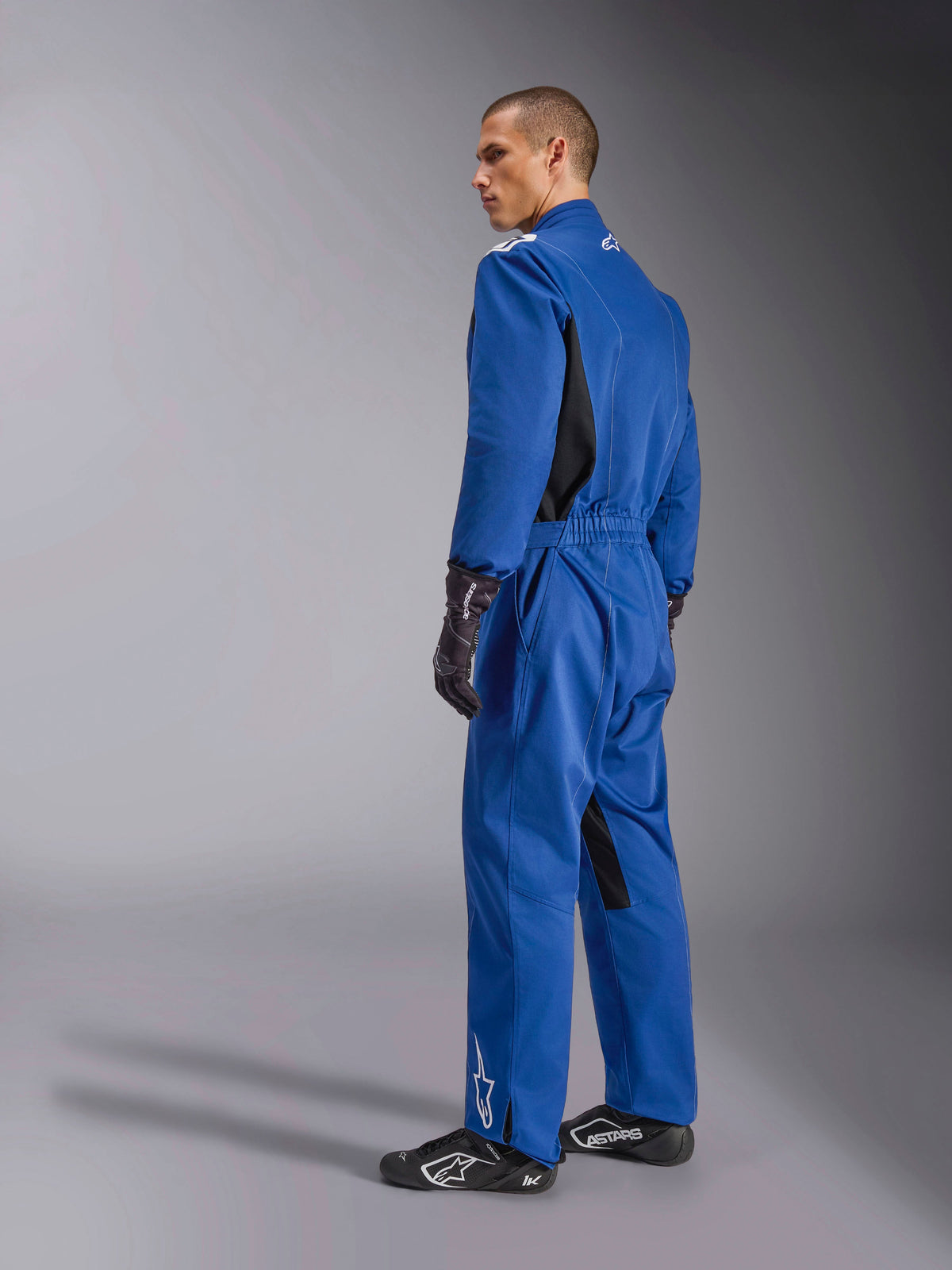 Combinaison Alpinestars Kart Indoor V2, combinaison de karting bleue, vue de dos et de côté, combinaison de course une pièce avec panneaux latéraux noirs et accents à l'intérieur des jambes, taille élastique et logos blancs, portée avec des gants de course et des chaussures noirs