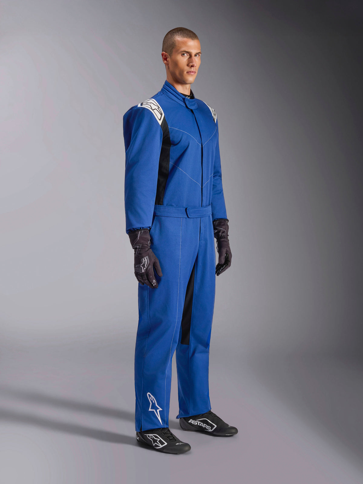 Combinaison Alpinestars Kart Indoor V2, combinaison de karting bleue, combinaison de course une pièce avec empiècements d'épaules et logos blancs, ceinture ajustable à la taille, logo blanc sur le bas de la jambe, portée par un mannequin avec gants et chaussures de course noirs, équipement de sport automobile pour le karting indoor