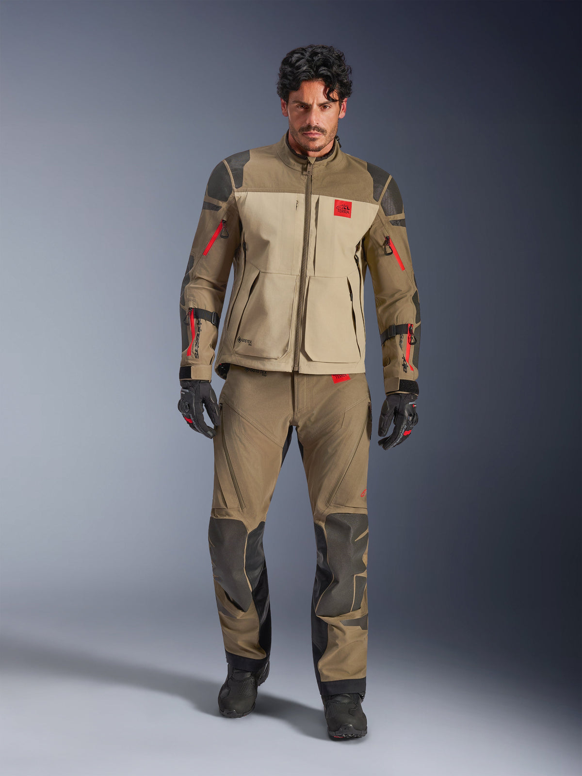 Alpinestars Nazca 3L GORE-TEX PRO Jacket, Walnut Rock Brown, adventure motorcycle riding jacket worn with matching pants and gloves, featuring red zipper accents, 'All Terra' chest logo, and rugged textile construction.
