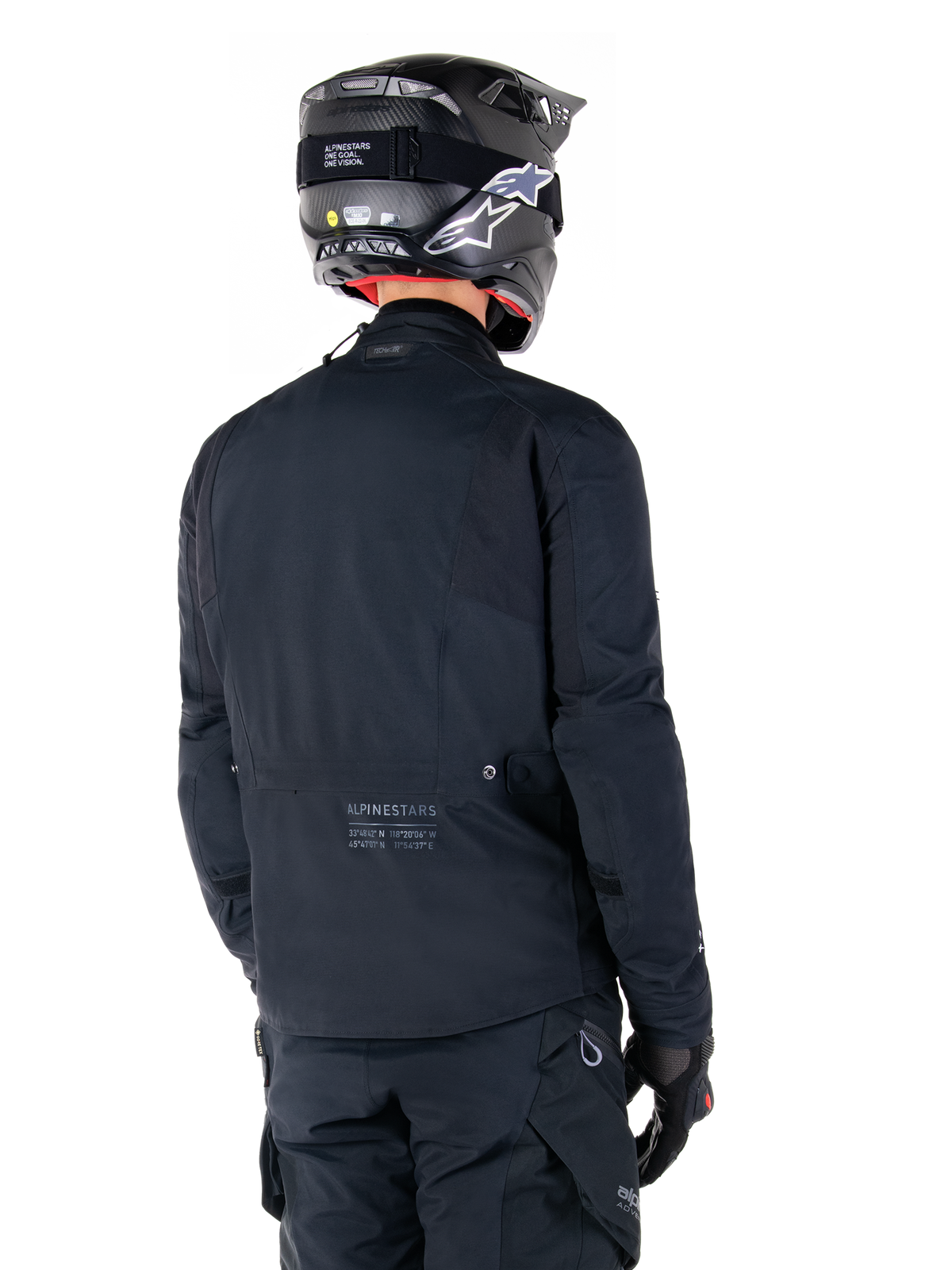 Alpinestars St-7 Prime GORE-TEX Jacket, Black, rear view of a rider wearing the jacket with a matching helmet and pants, featuring Alpinestars branding and geographic coordinates on the lower back.