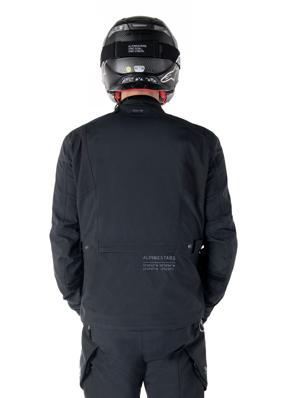 Alpinestars St-7 Prime GORE-TEX Jacket, Black, rear view of a rider, featuring subtle logo and geographic coordinates on the lower back, worn with a matching black helmet and pants, sleek and technical motorcycling apparel
