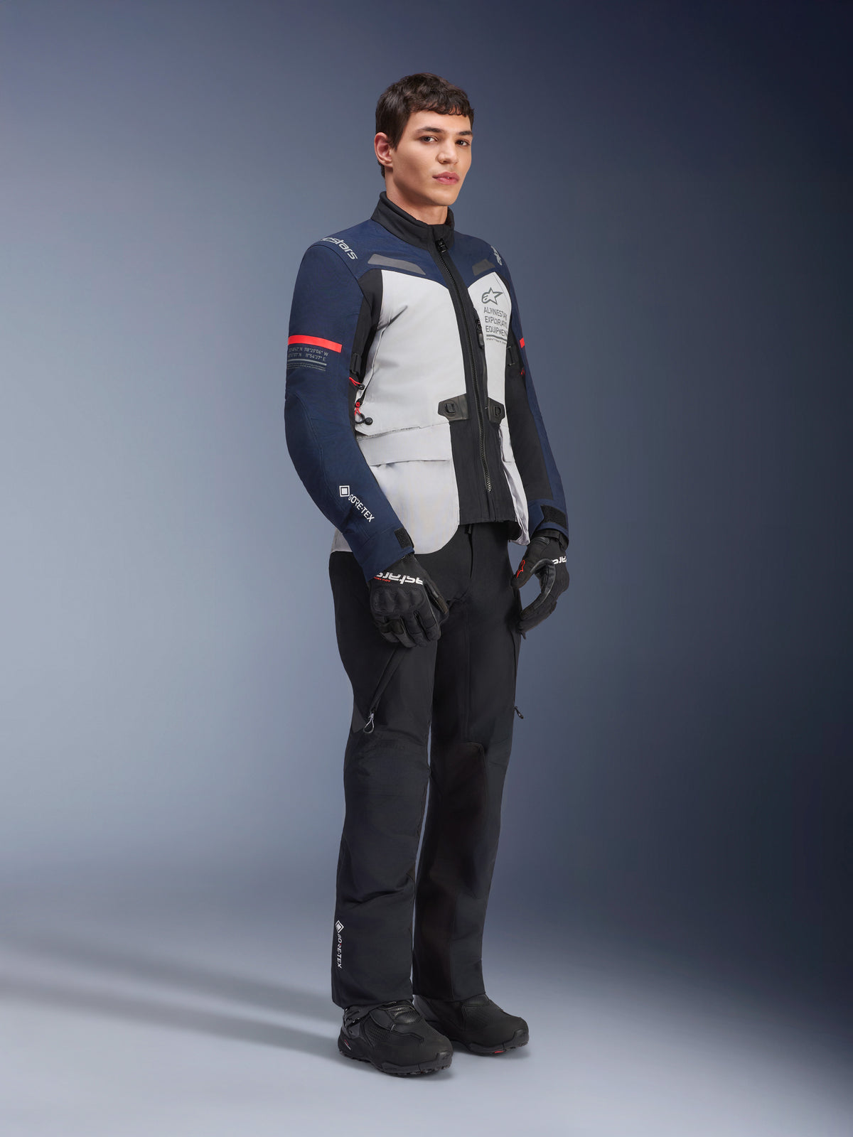 Veste Alpinestars ST-7 2L GORE-TEX en coloris Gris Glace, Bleu Foncé et Noir avec des accents rouges, portée par un mannequin homme avec pantalon et gants de moto noirs, équipement de moto d'aventure touring professionnel